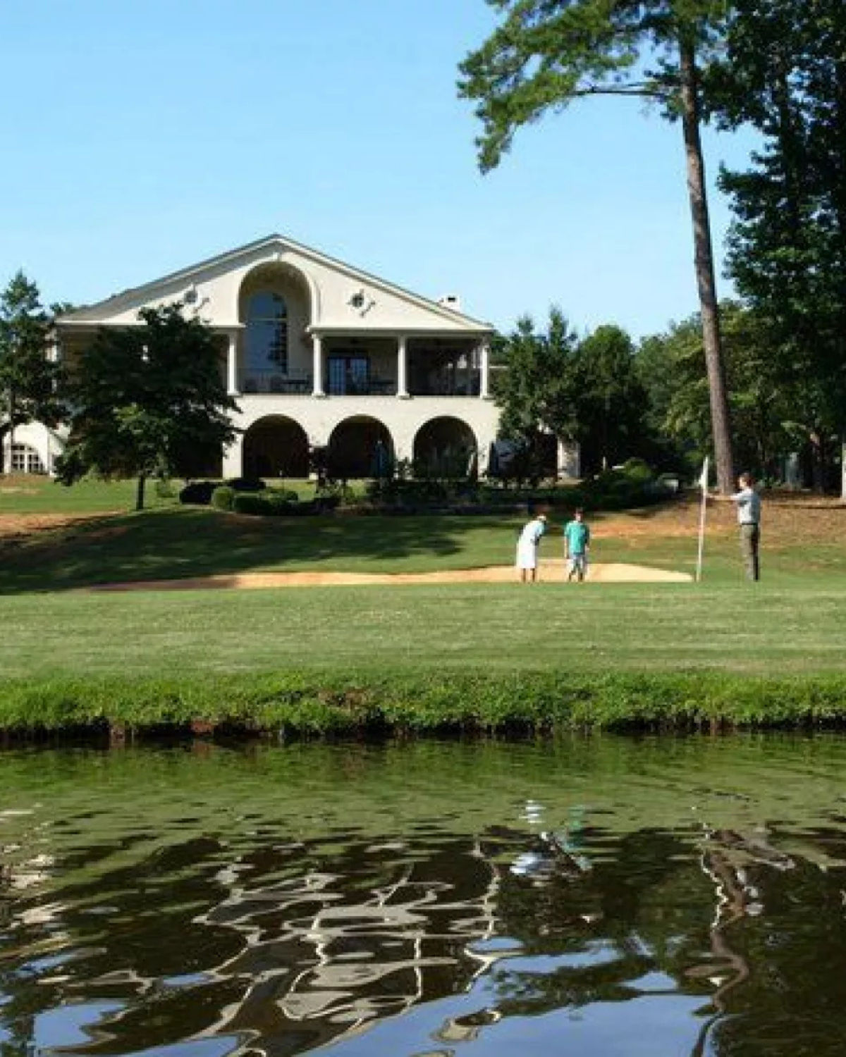 Lakeside view of The Club at Shoal Creek in Griffin, Georgia, featuring golfers putting on a green beside calm water, with a large balconied clubhouse