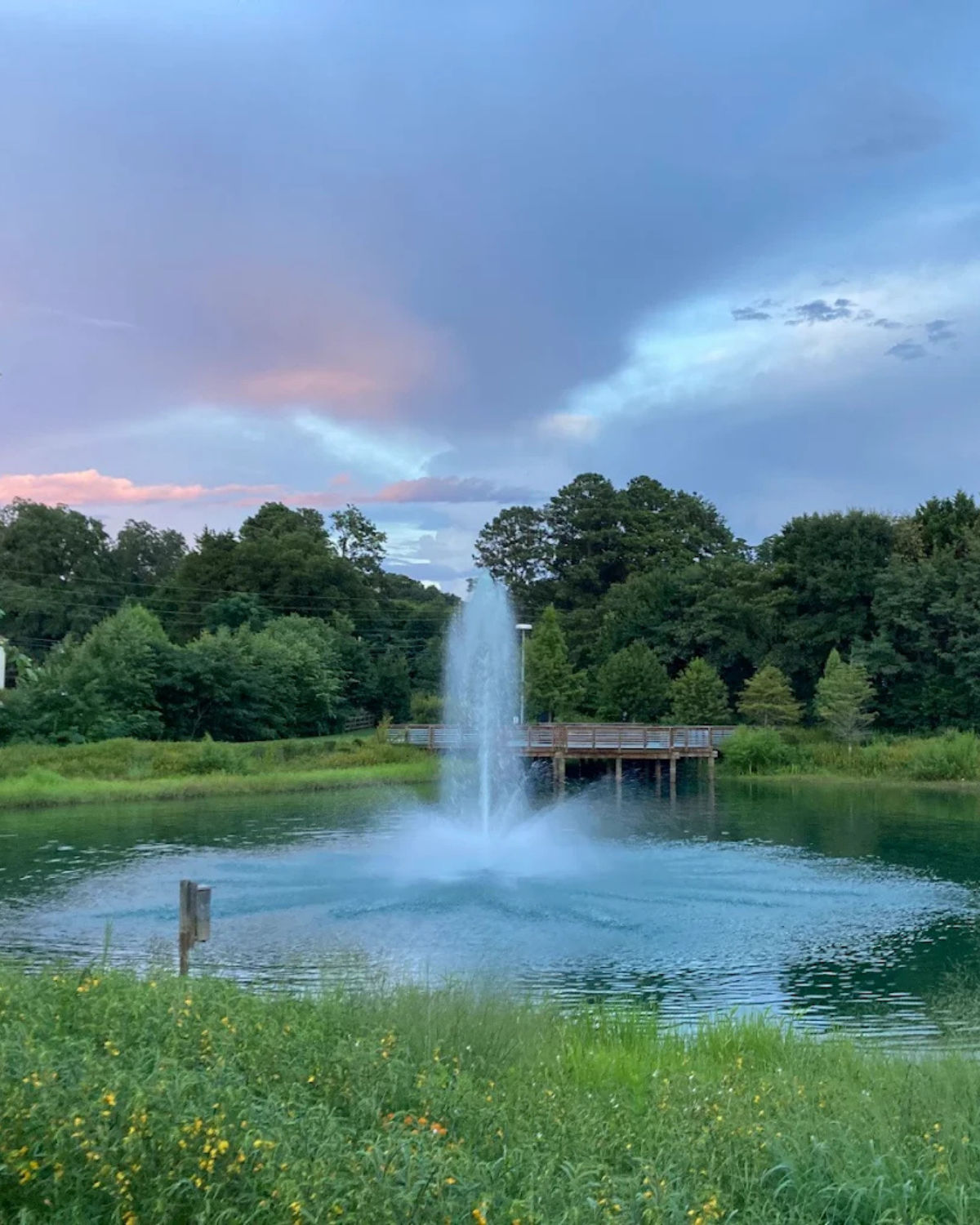 Sunset view of the pond at Big Springs Park in McDonough, Georgia, with a tall fountain spraying into the water, surrounded by grassy wildflowers and trees, with a wooden boardwalk overlook in the background