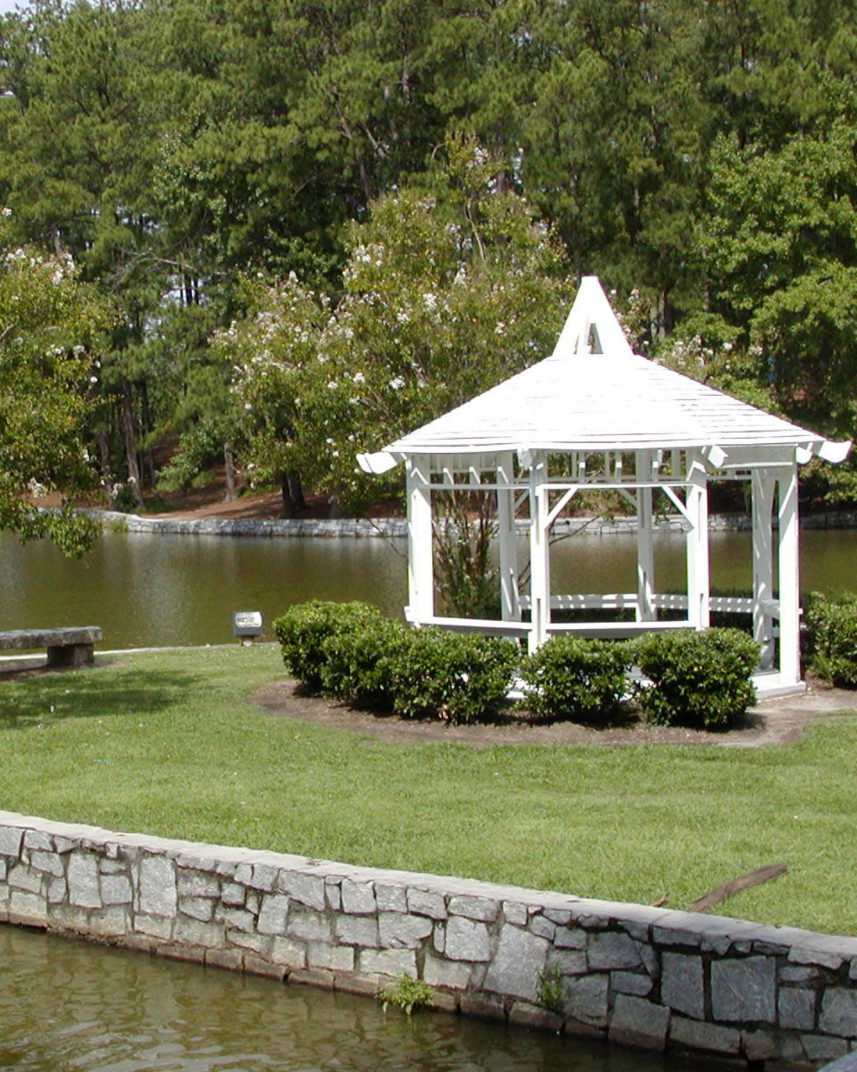 White gazebo by the pond in Briscoe Park in Snellville, Georgia, surrounded by neatly trimmed shrubs, grassy banks, and tall trees reflected in the calm water.