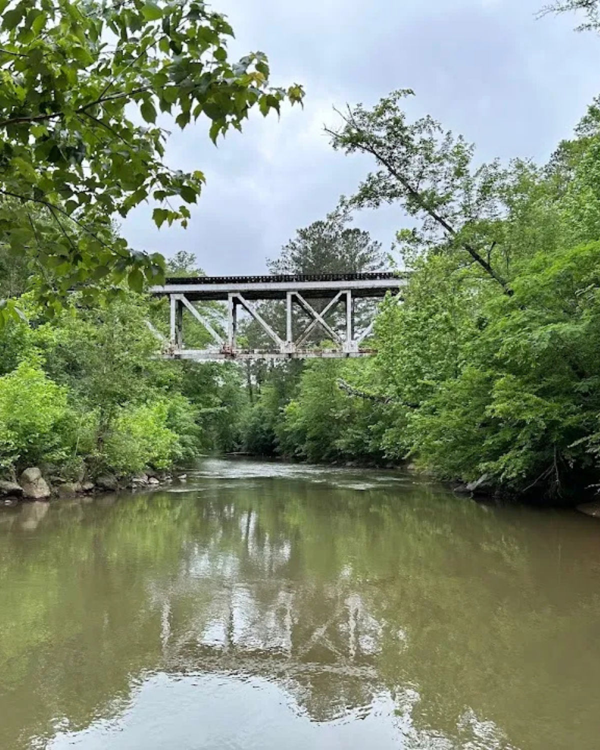 River view at Olde Rope Mill Park in Woodstock, Georgia, showing a stretch of the Little River and green foliage, with an elevated white railroad bridge
