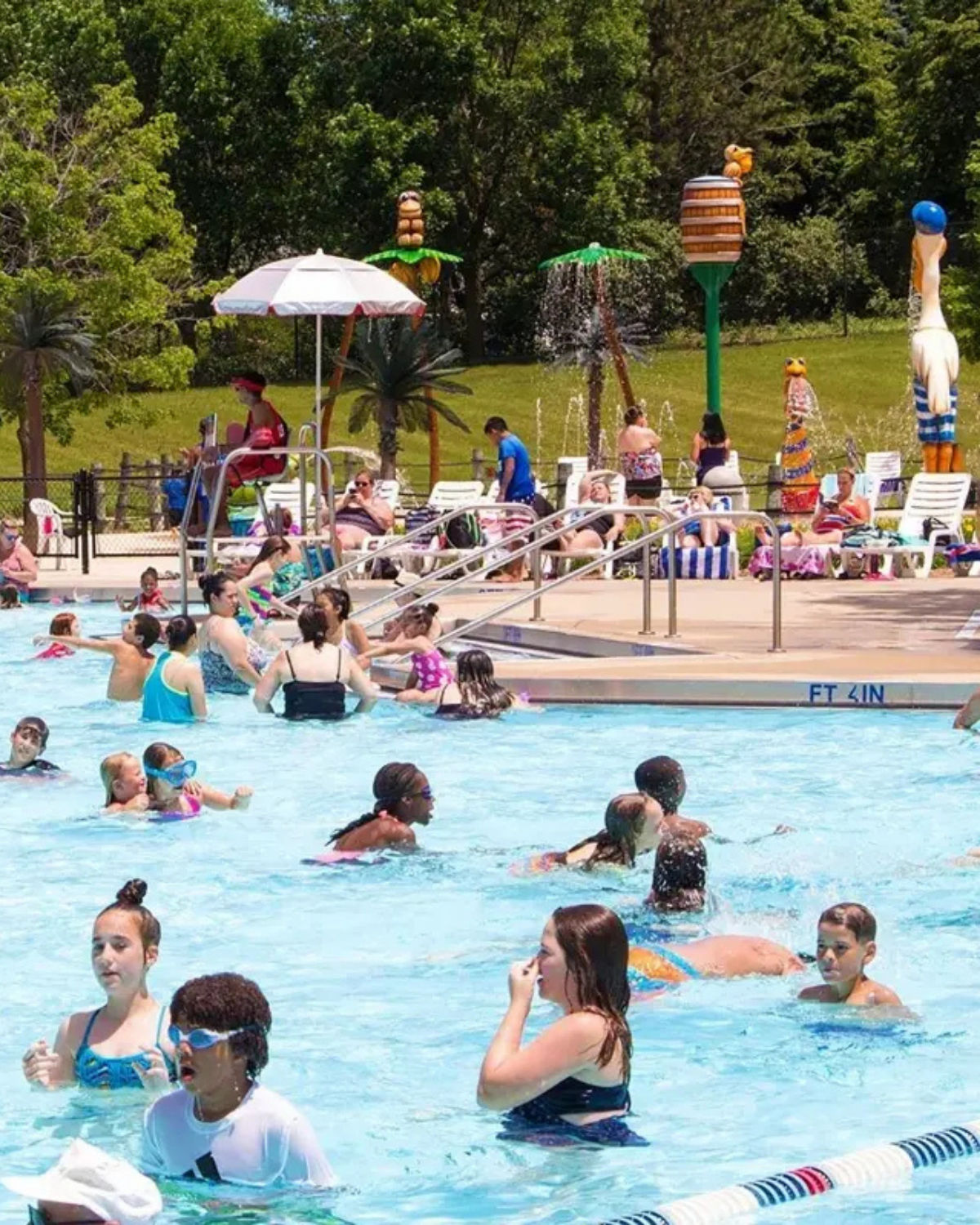 Crowded outdoor pool at Pelican Harbor Water Park in Bolingbrook, Illinois, with kids and families swimming while lifeguards watch from poolside chairs and colorful spray features decorate the background lawn.