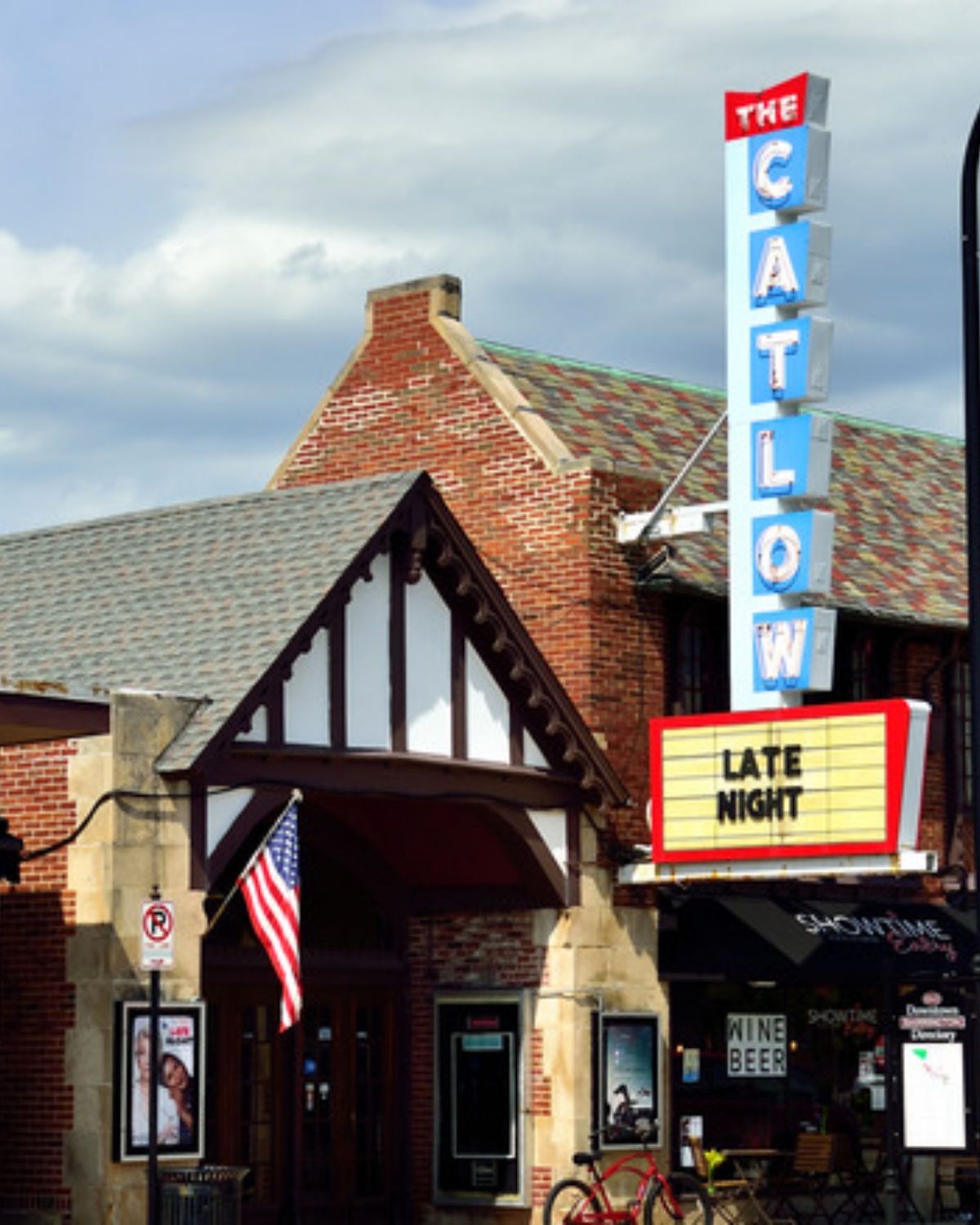 Historic brick theater building with a Tudor-style timber gable entrance and a tall vertical neon sign reading ÒThe Catlow.Ó The marquee reads ÒLate Night,Ó and an American flag hangs near the entrance under a partly cloudy