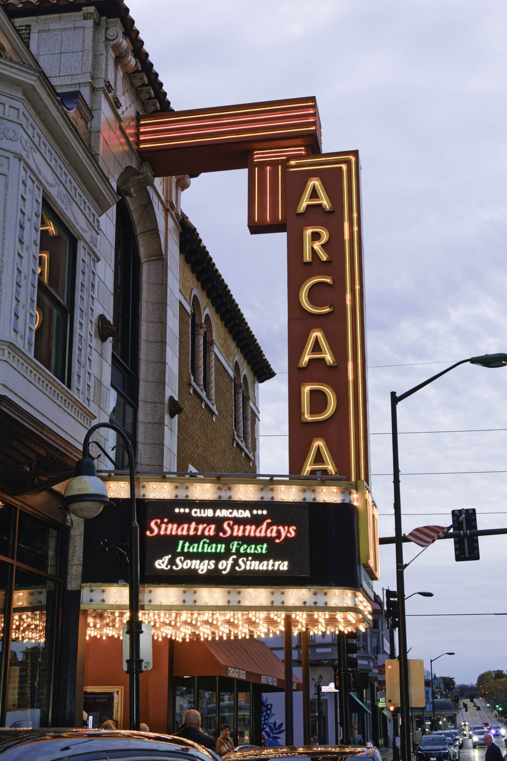 Close-up view of the historic Arcada Theatre marquee near Carol Stream, Illinois, glowing with vibrant neon lights above a lively suburban downtown street lined with restaurants, storefronts, and evening traffic.
