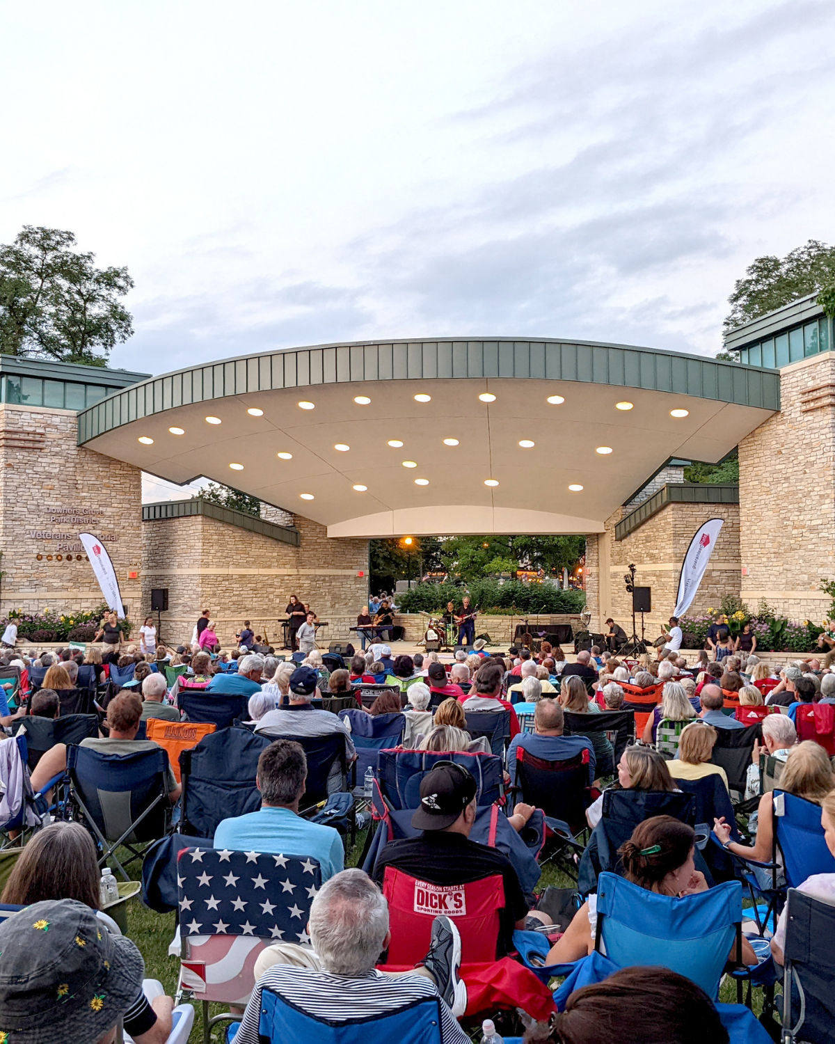 Evening concert at the Veterans Memorial Pavilion in Fishel Park in Downers Grove, Illinois, with a large crowd in lawn chairs facing the modern stone-and-metal bandshell as musicians perform on stage.