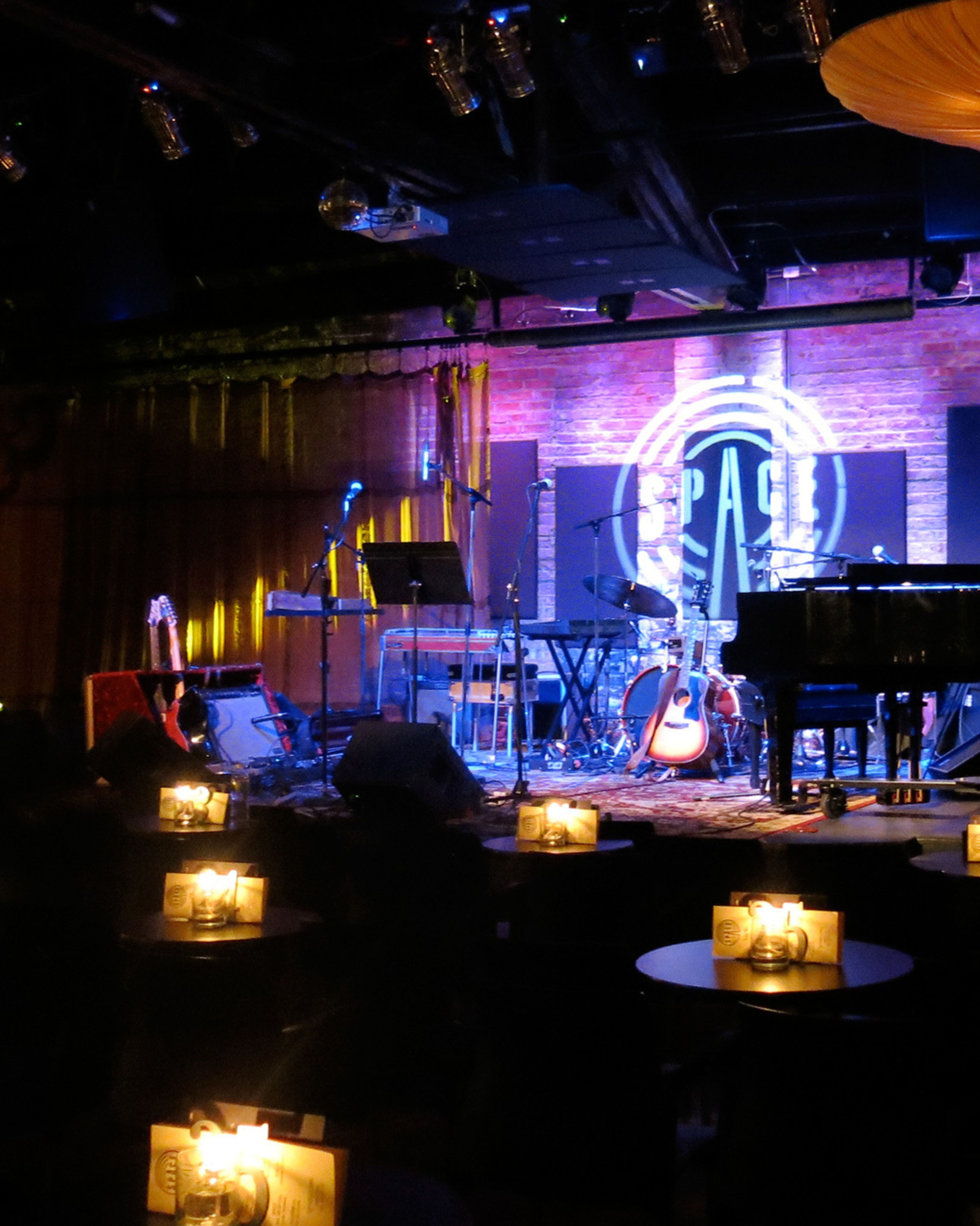 Interior view of SPACE Evanston, a cozy live music venue near downtown Evanston, Illinois, featuring a softly lit stage with instruments, microphones, neon signage, and intimate candlelit seating for live performances and concerts.