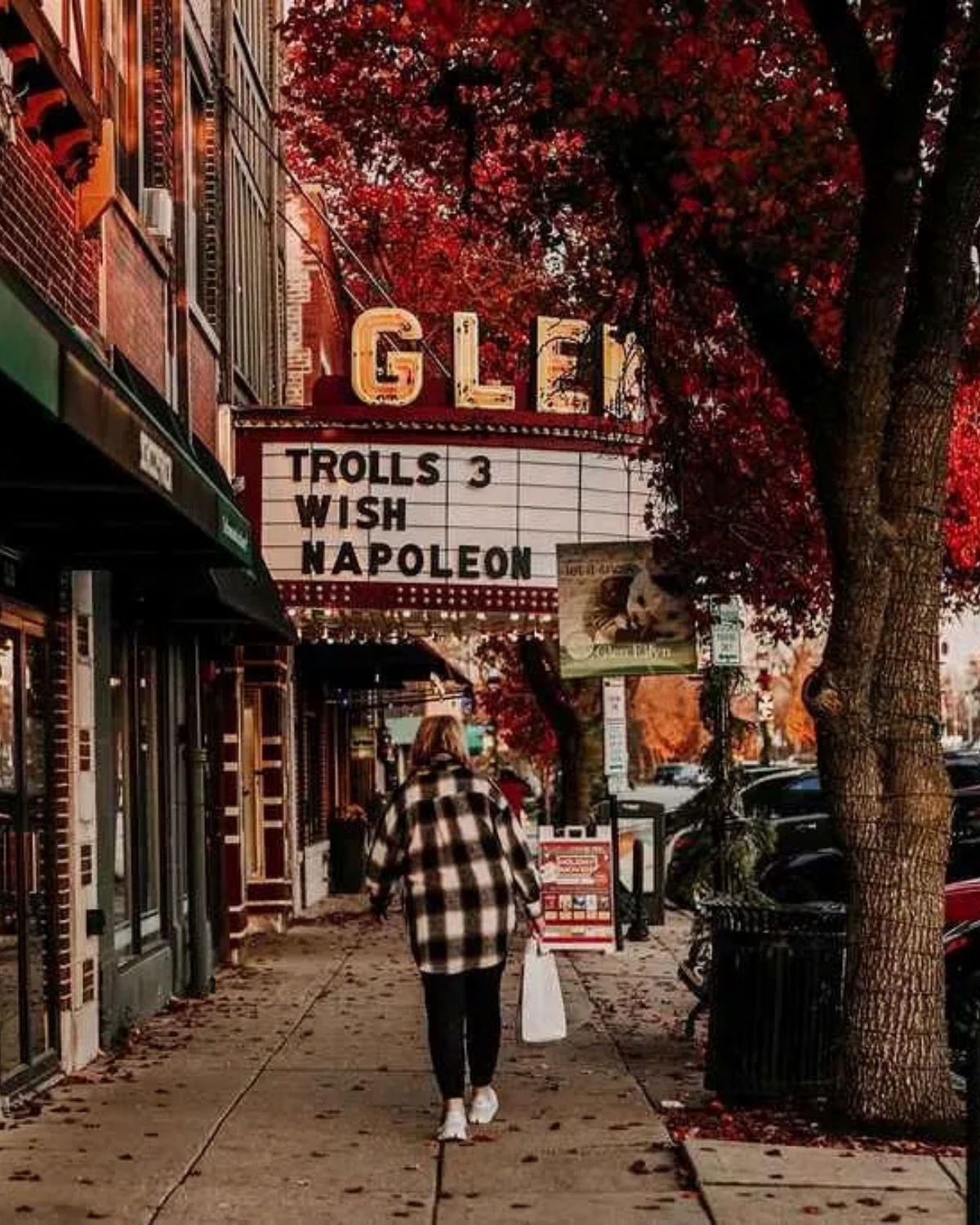 Pedestrian walking past the Glen Art Theatre in downtown Glen Ellyn, Illinois, with the vintage “GLEN” marquee listing films like “Trolls 3,” “Wish,” and “Napoleon” above a tree-lined sidewalk covered in autumn leaves.