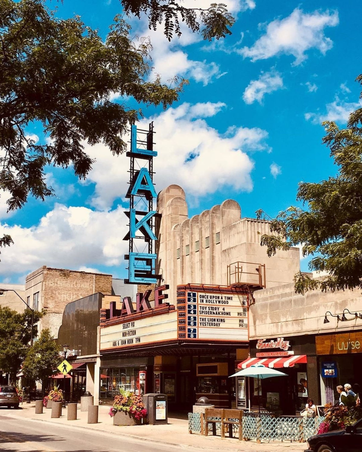Street-level view of the Classic Cinemas Lake Theatre on Lake Street in Oak Park, Illinois, featuring its tall blue vertical ŇLAKEÓ neon sign and retro signage above the sidewalk