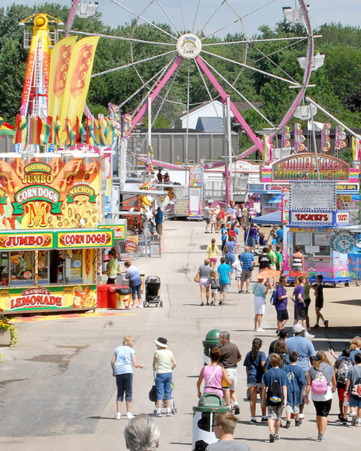 Busy summer day at the DuPage County Fair in Wheaton, Illinois, with crowds walking between bright food stands selling corn dogs and lemonade, and a large pink-and-white Ferris wheel and rides towering in the background.