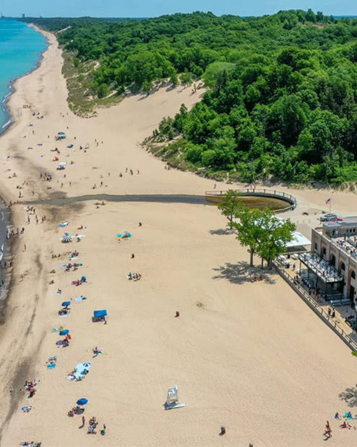 Aerial view of a packed sandy beach along Lake Michigan with swimmers, sunbathers, and a lakeside pavilion on a sunny summer day in Gary, Indiana