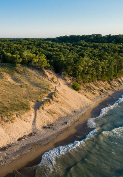 Aerial coastal view near Hammond, Indiana, featuring sandy dunes, forest-lined shoreline, rolling waves along Lake Michigan, and scenic hiking trails stretching through a peaceful natural landscape.