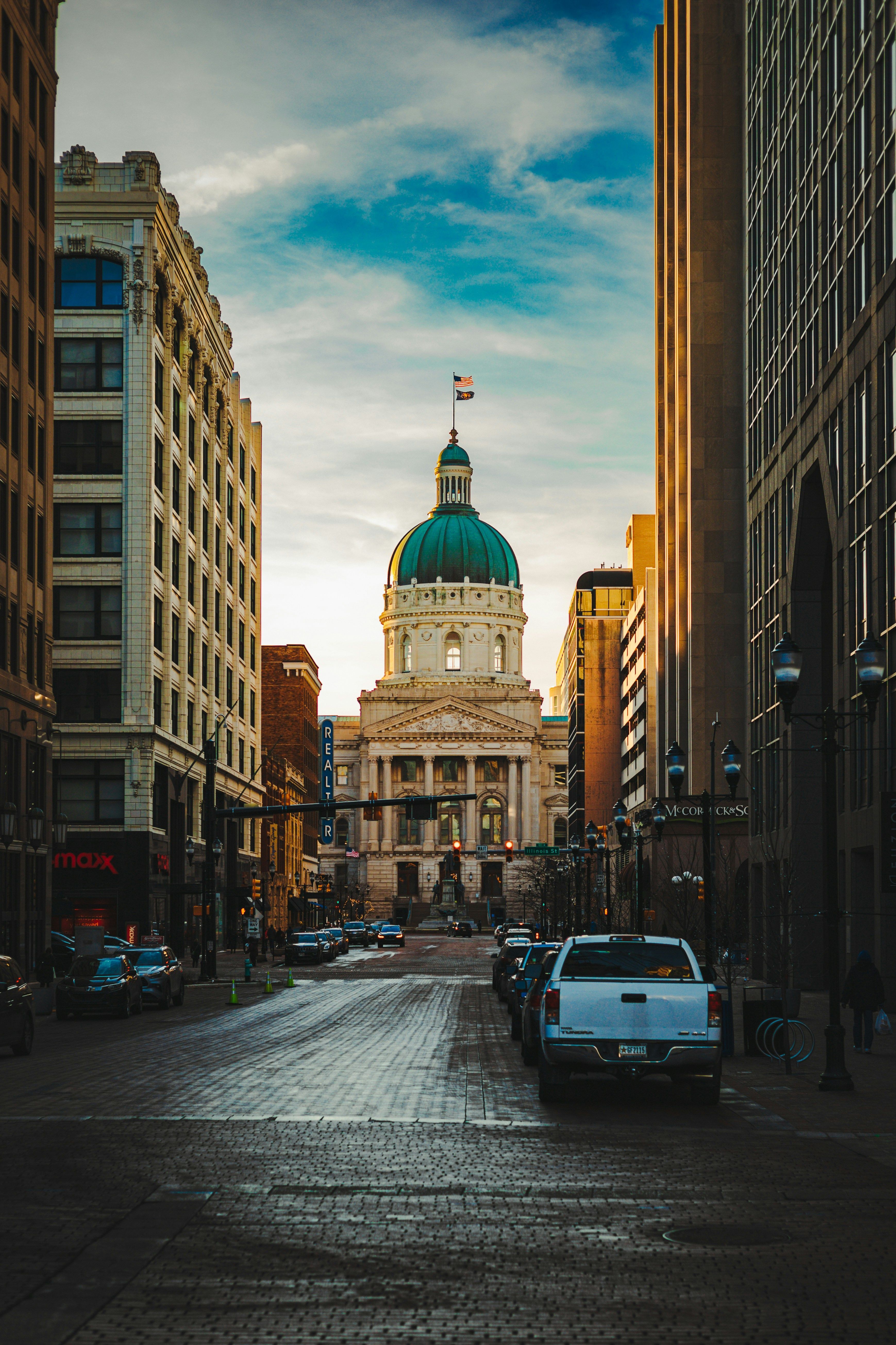 Street-level view of downtown Indianapolis with the Indiana State Capitol dome rising between modern buildings under a dramatic cloudy sky