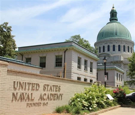View of the United States Naval Academy in Annapolis, Maryland, featuring the entrance sign in the foreground and the iconic domed chapel rising behind it, surrounded by manicured greenery and historic campus architecture.