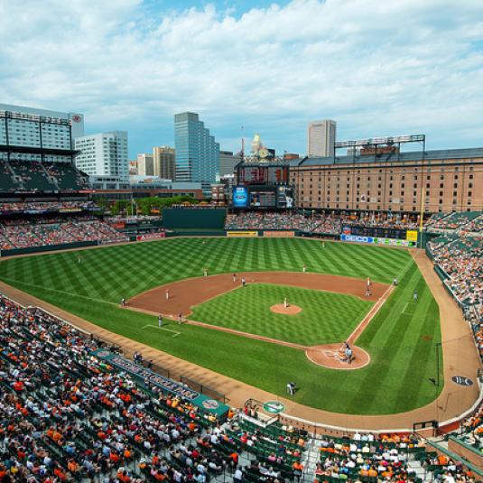 Wide view of a baseball game in progress at Oriole Park at Camden Yards in Baltimore, Maryland, with the city skyline and historic warehouse visible beyond the outfield and fans filling the stadium seats.