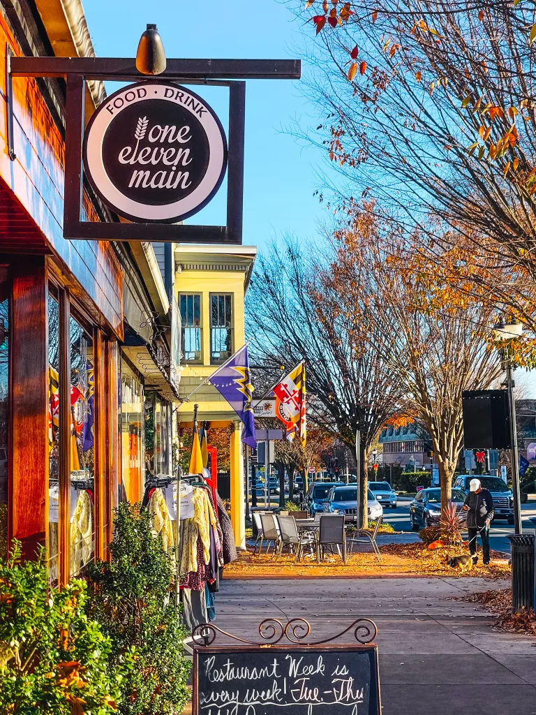 Sidewalk view of downtown Bel Air, Maryland, featuring the storefront of One Eleven Main restaurant, outdoor seating, local shops, Maryland flags, and tree-lined streets with autumn foliage.