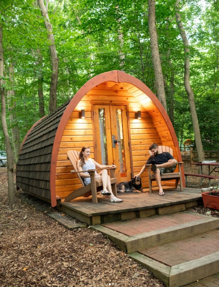 Wooden glamping cabin at Cherry Hill Park in Beltsville, Maryland, featuring a cozy curved-roof cabin with a front deck, Adirondack chairs, towering trees, and guests relaxing with their dog in a peaceful forest setting.