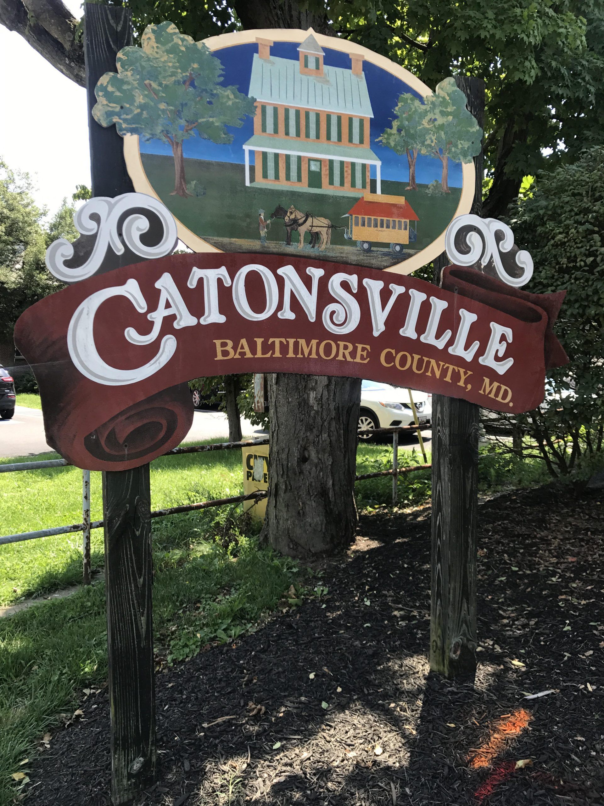 Welcome sign for Catonsville, Maryland in Baltimore County, featuring a decorative illustration of a historic home and carriage, surrounded by trees in a suburban neighborhood setting.