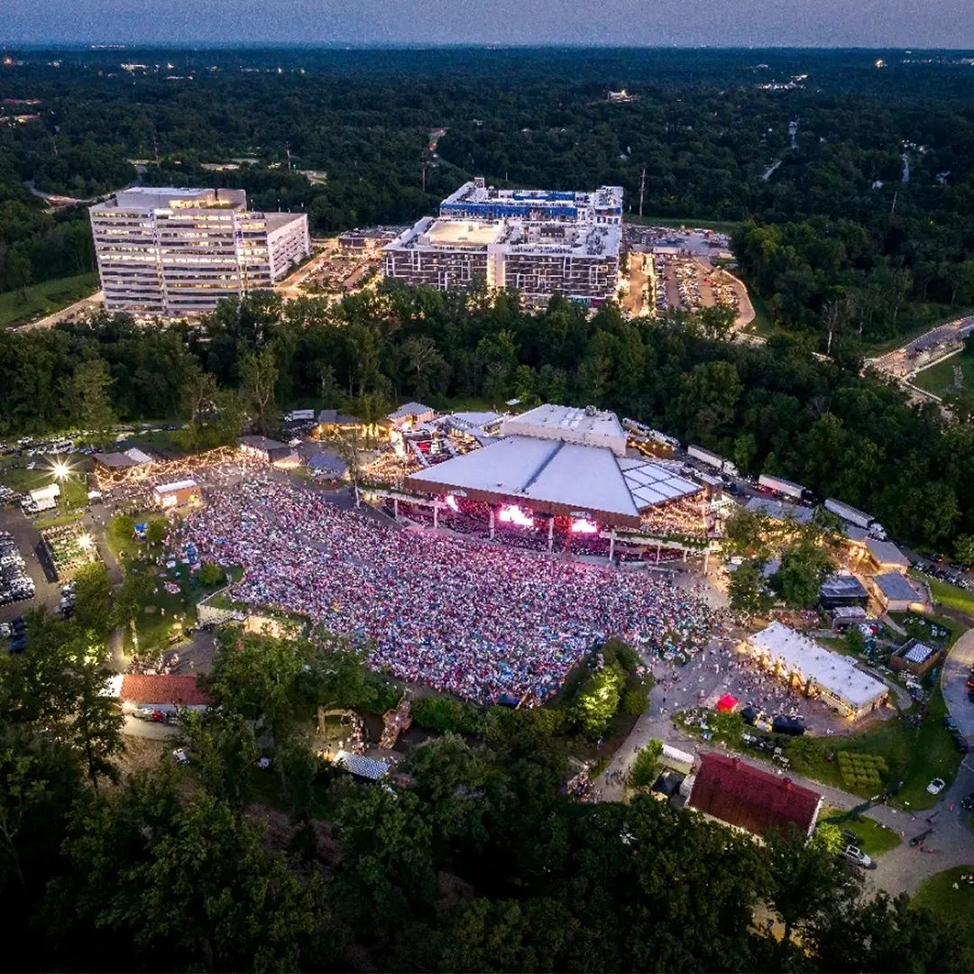Aerial view of a large outdoor concert at Merriweather Post Pavilion in Columbia, Maryland at dusk, featuring a packed crowd gathered around the open-air stage, surrounding wooded areas, nearby buildings, and event lighting illuminating the venue as the sun sets.
