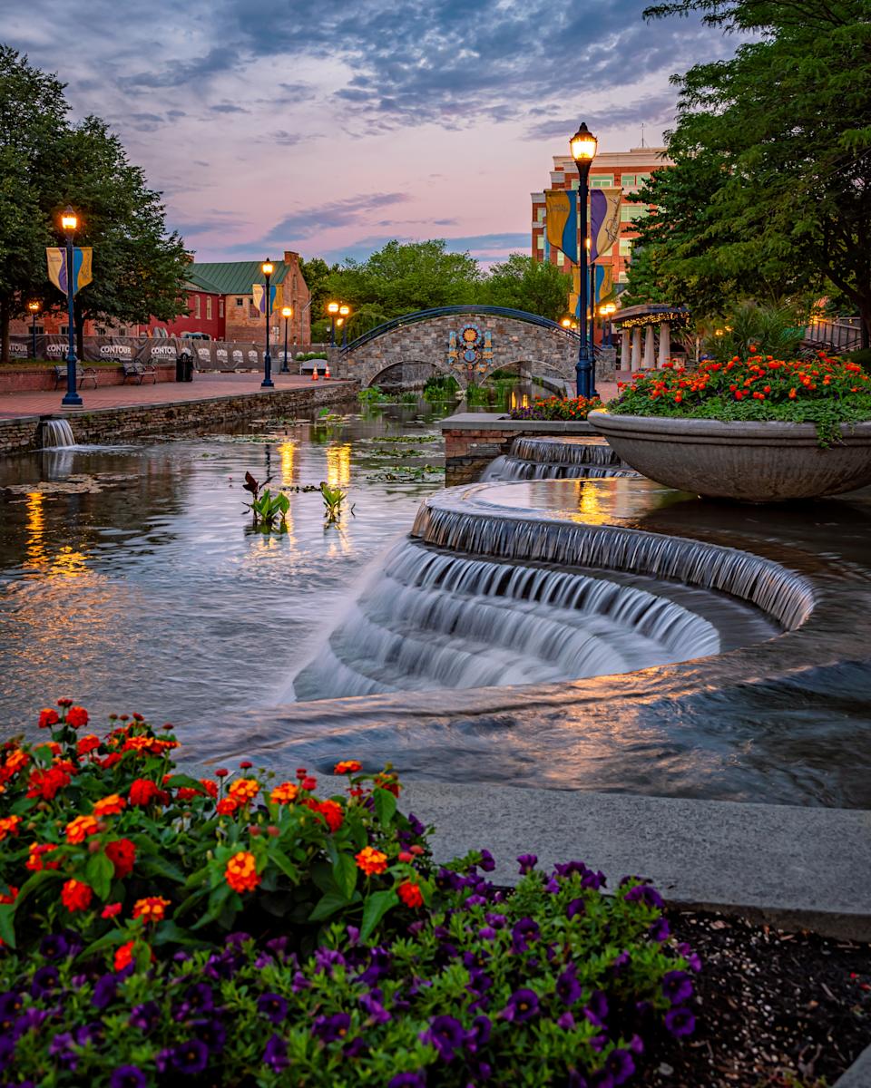 Carroll Creek Park in downtown Frederick, Maryland, at dusk. Cascading water features flow through the landscaped urban park, surrounded by colorful flowers, decorative bridges, glowing street lamps, and brick walkways that create a picturesque waterfront setting in the heart of the city.