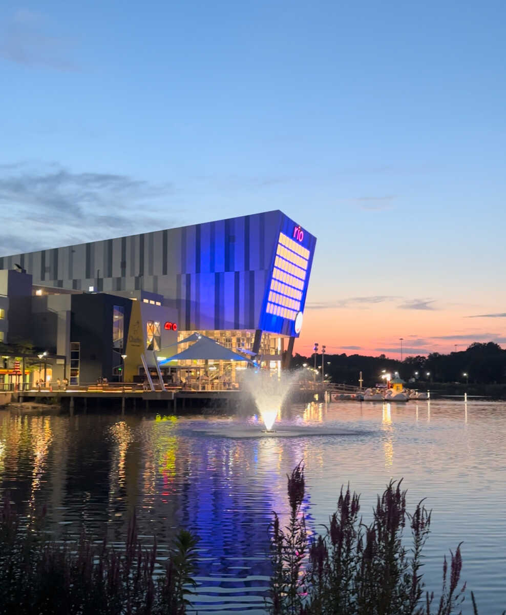 Evening view of RIO Lakefront in Gaithersburg, Maryland, featuring illuminated modern buildings, waterfront dining, a lit fountain in the lake, and reflections across the water at sunset.