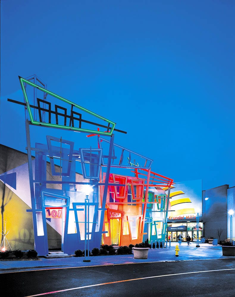 Colorful exterior of the Arundel Mills shopping area in Glen Burnie, Maryland at dusk, featuring bold geometric architectural designs, bright neon lighting, and the entrance to the mall surrounded by a lively retail and entertainment setting.