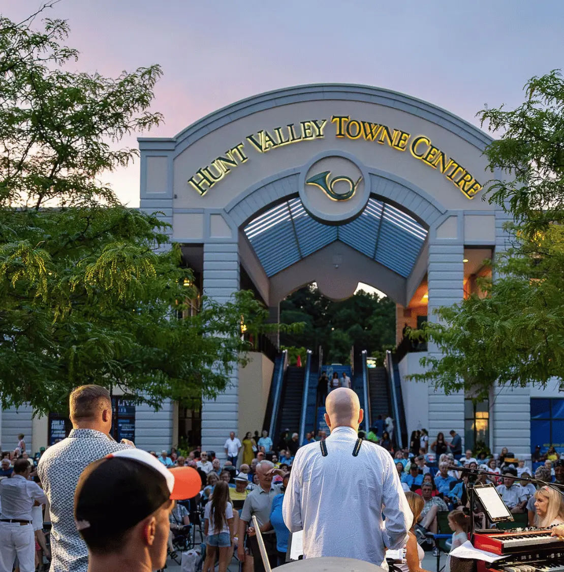 Entrance to Hunt Valley Towne Centre in Hunt Valley, Maryland, featuring the shopping center's arched gateway, illuminated signage, outdoor escalators, and a crowd gathered for a live community event.