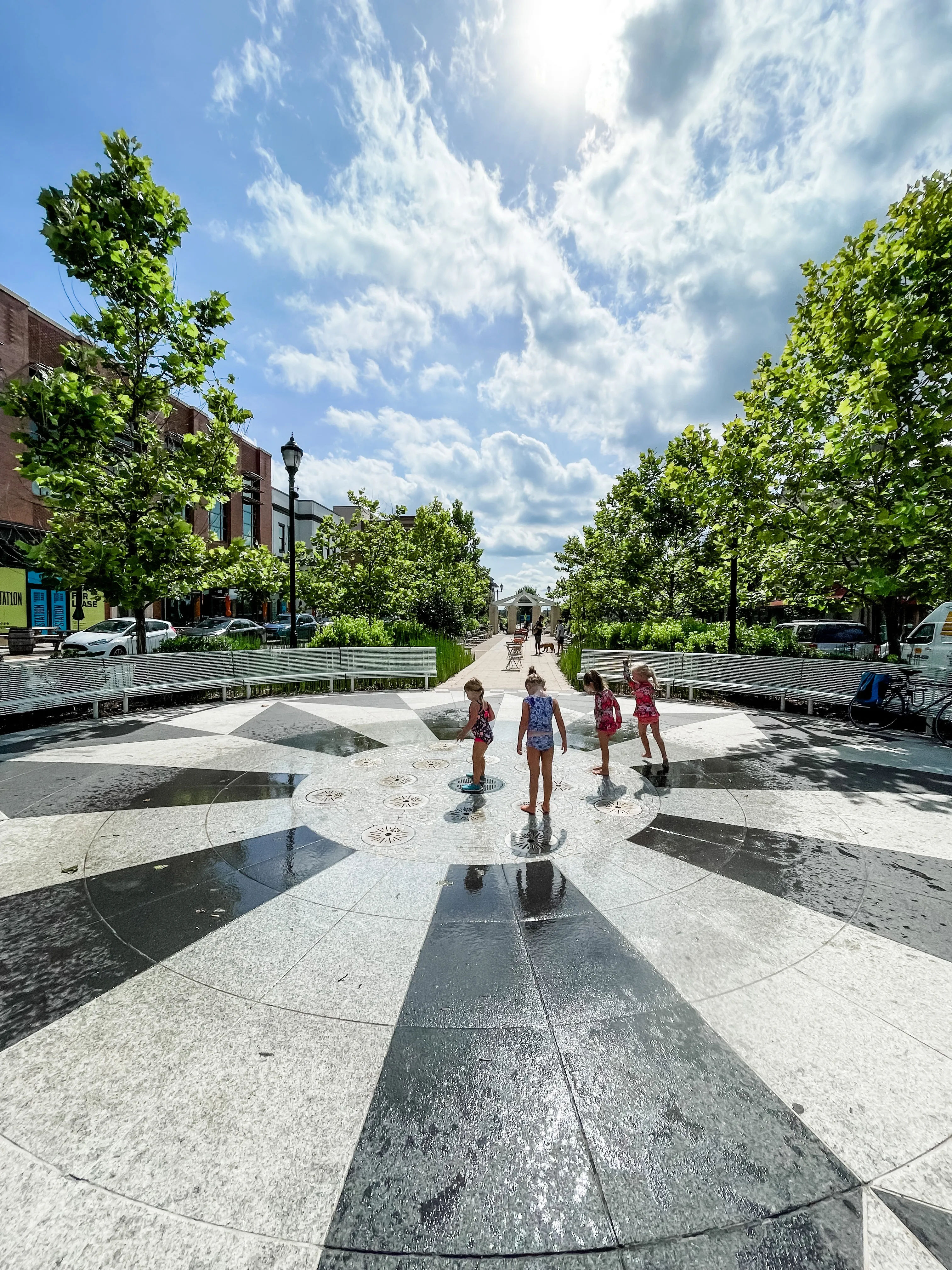 Children playing in a splash pad at a Hyattsville, Maryland community plaza, featuring modern landscaping, tree-lined walkways, and a vibrant outdoor gathering space on a sunny day.