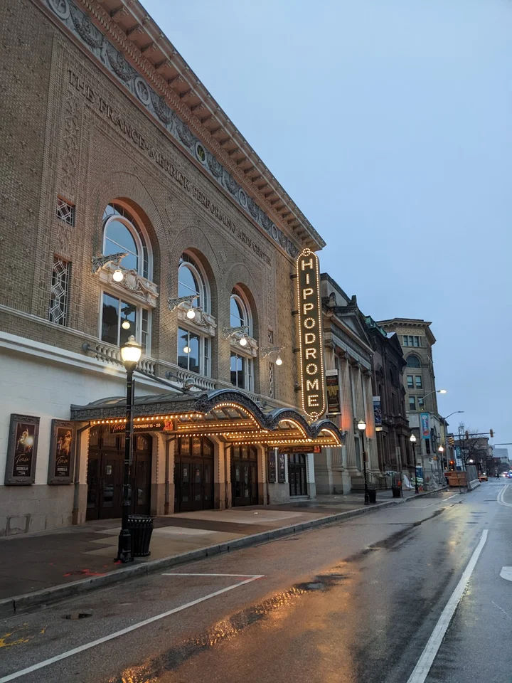 Street-level view of the historic Hippodrome Theatre in downtown Baltimore near Pikesville, Maryland, featuring the illuminated vertical marquee, ornate brick façade, glowing entrance lights, and a rain-soaked city street at dusk.