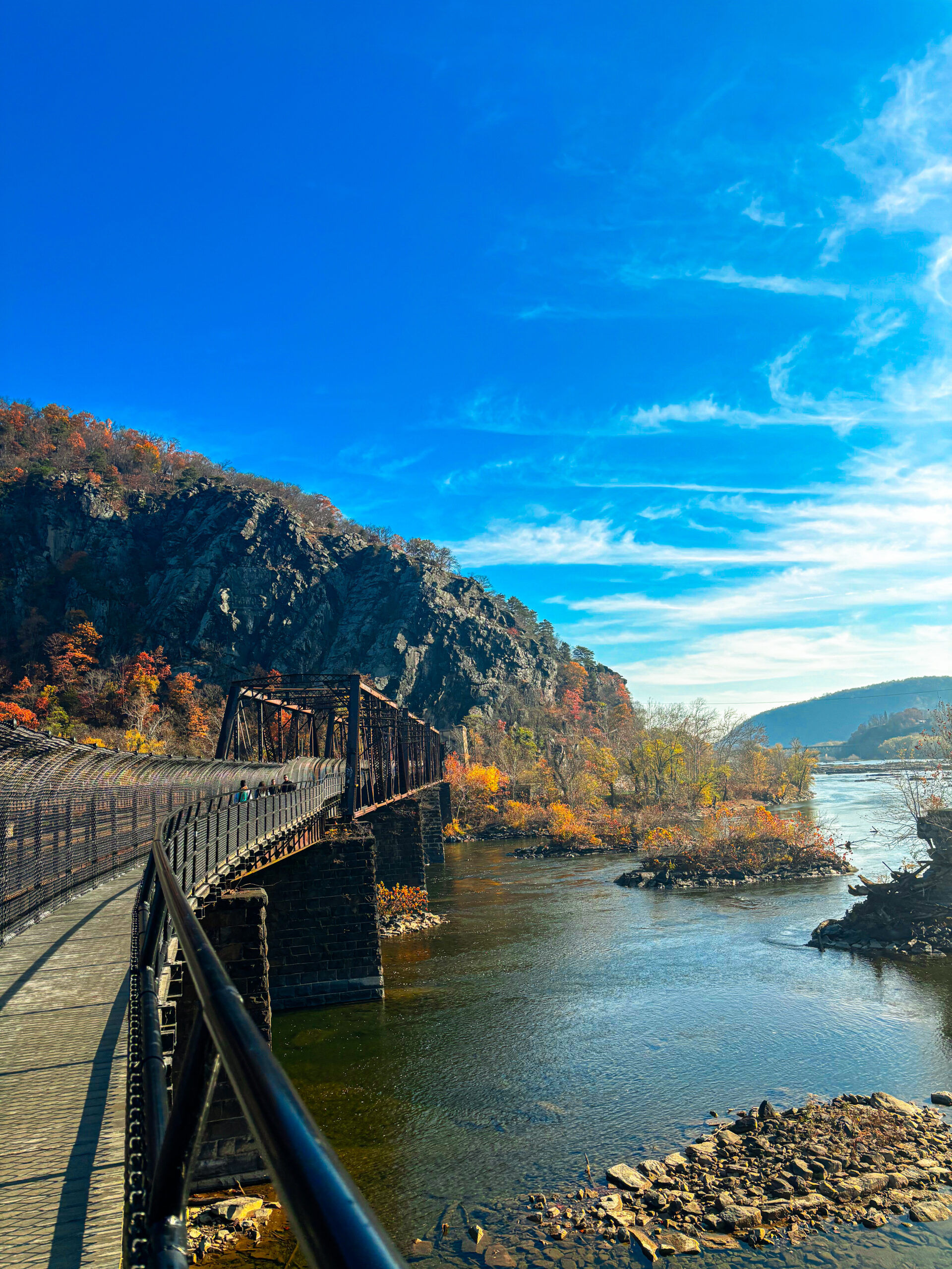 Scenic view along the C&O Canal Towpath near Potomac, MD featuring a pedestrian bridge over the Potomac River, surrounded by rocky cliffs and fall foliage under a bright blue sky