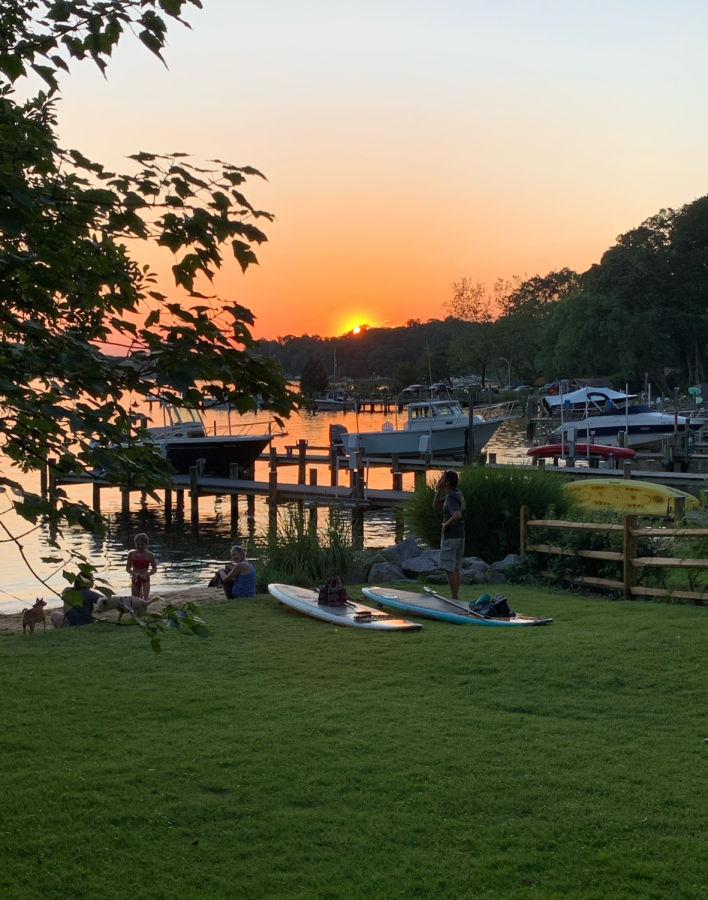 Sunset waterfront scene in Severna Park, Maryland featuring boats docked along the shoreline, people relaxing on the grass, and paddleboards near the water with warm evening light.