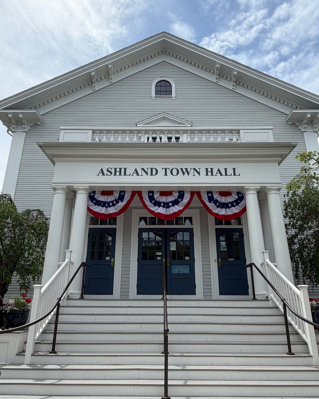 Front entrance of Ashland Town Hall in Ashland, Massachusetts, featuring a classic white New England civic building with tall columns, blue doors, patriotic red-white-and-blue bunting, and a grand staircase beneath a bright cloudy sky.