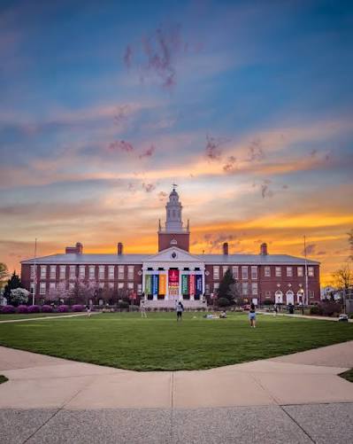 A sunset view of the main lawn and historic administration building at Bridgewater State University in Bridgewater, Massachusetts, with students gathered on the campus green beneath a colorful evening sky.