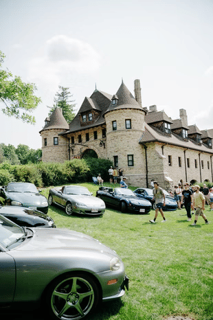 Hammond Castle Museum in Brookline, Massachusetts, featuring a stone castle-style building with turreted towers, surrounded by classic sports cars and visitors attending an outdoor community event.