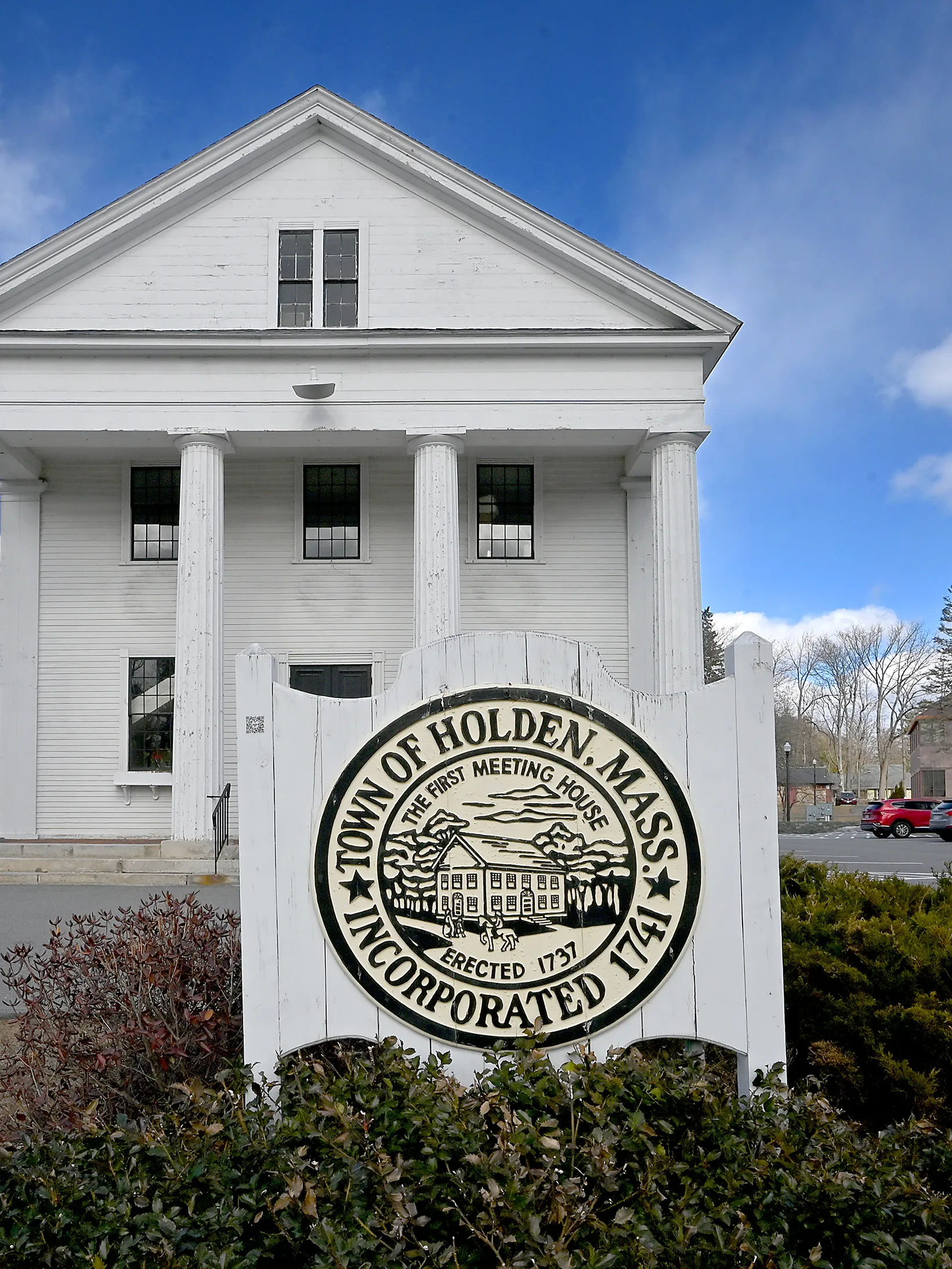 Historic white meeting house in Holden, Massachusetts with tall columns and a town seal sign marking its incorporation in 1741 under a clear blue sky