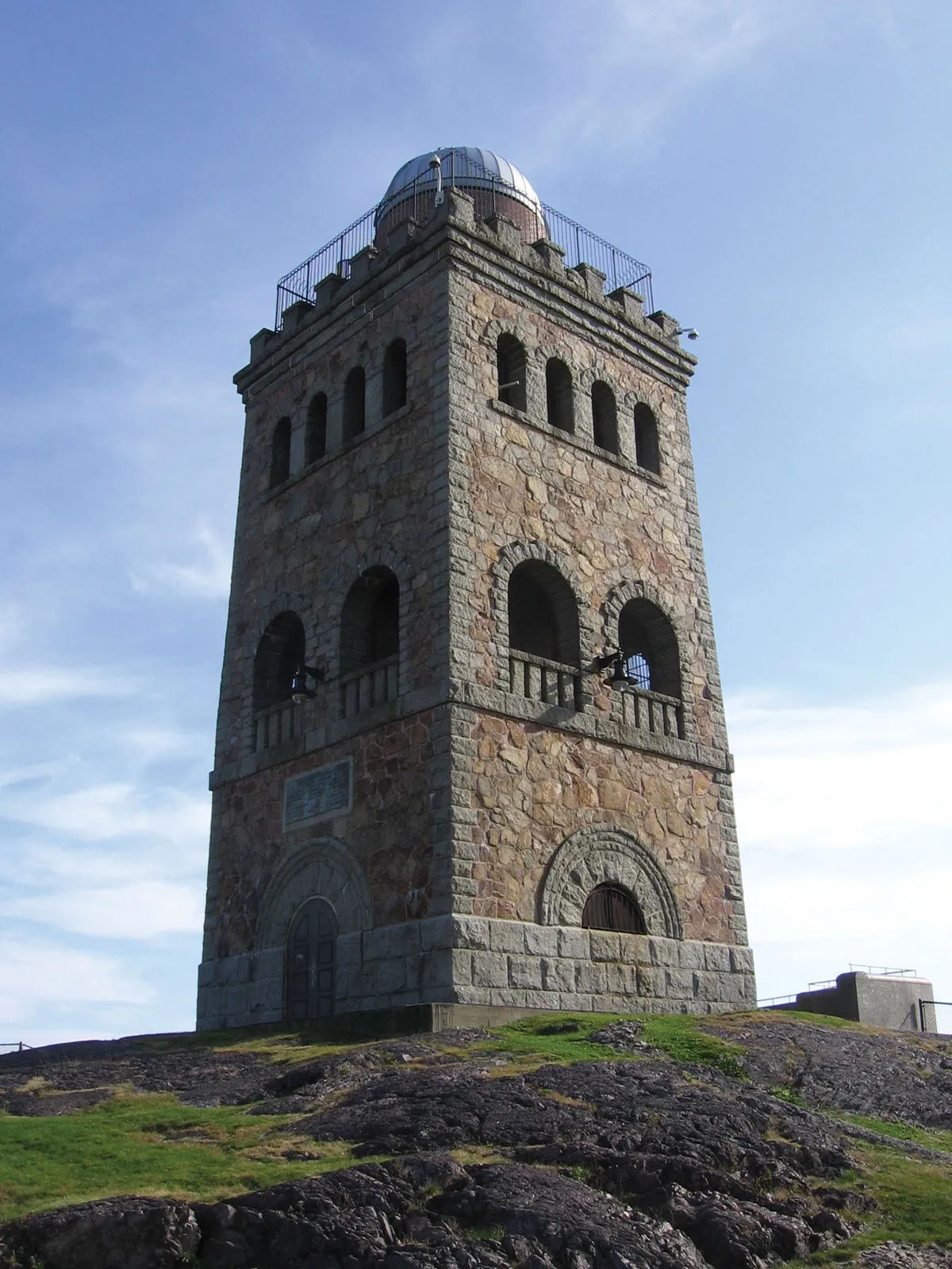 Stone observation tower at High Rock Tower Reservation in Lynn, Massachusetts, featuring the historic granite structure overlooking rocky terrain beneath a bright blue sky.