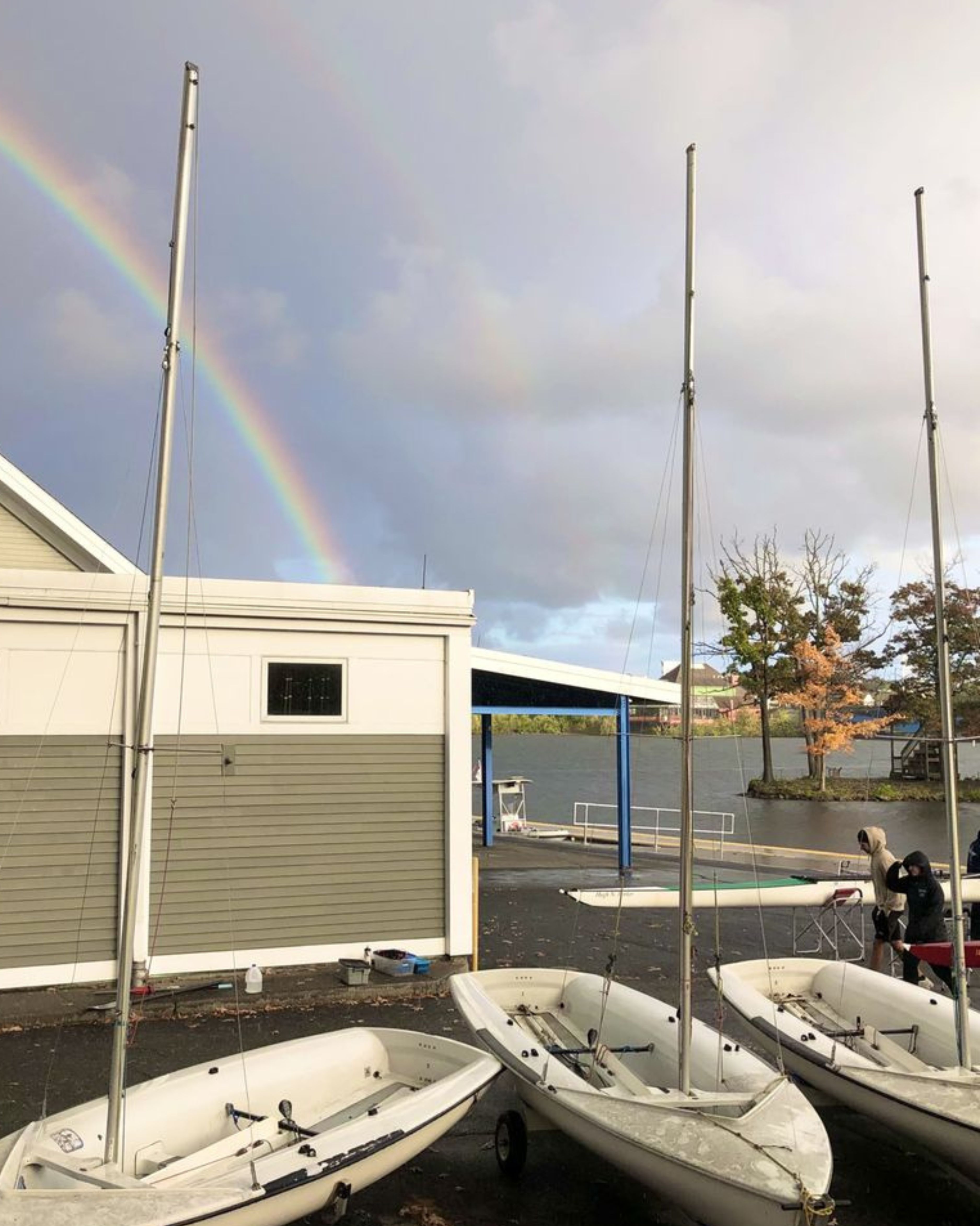 Small sailboats lined up outside a boating facility in Millbury, MA, as a rainbow stretches across a cloudy sky above the water following a passing rain shower.