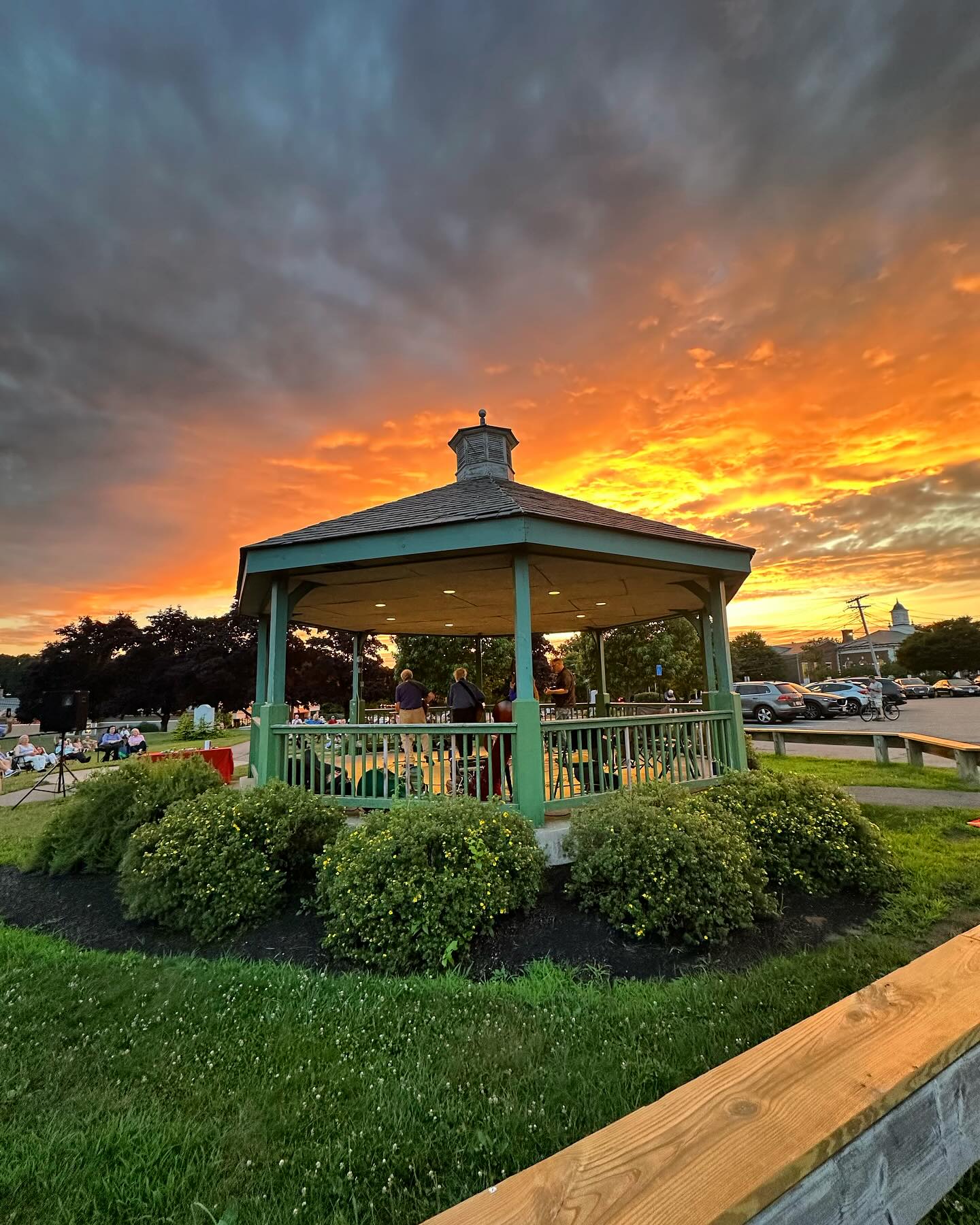 Sunset view of a community gazebo in Needham, Massachusetts, featuring people gathered for an outdoor event beneath a dramatic orange and blue evening sky, surrounded by green landscaping, local park space, and small-town New England charm.