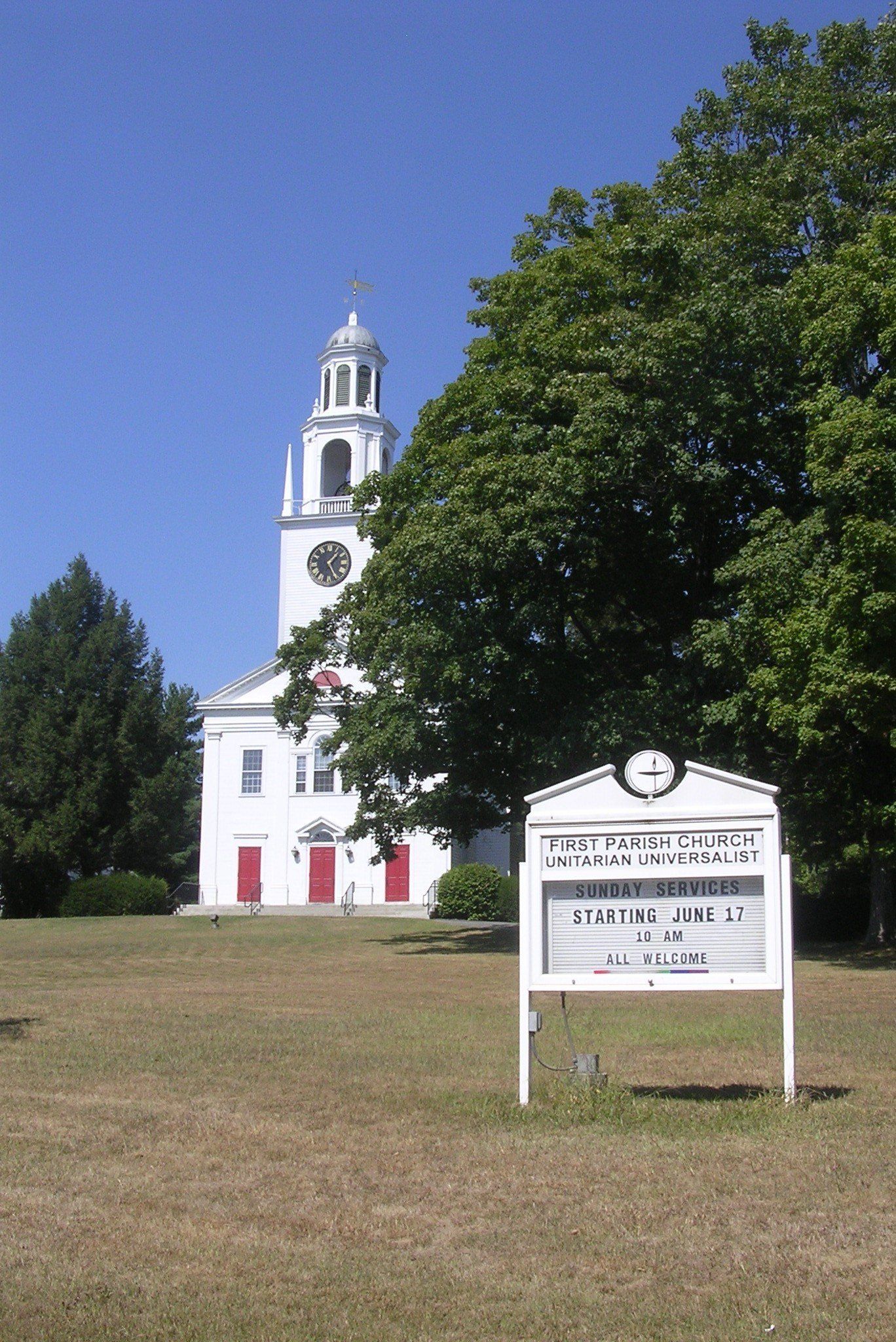 Exterior view of First Parish Church in Northborough, Massachusetts, featuring the historic white church building with its clocktower steeple, open lawn, and surrounding mature trees under a clear blue sky.