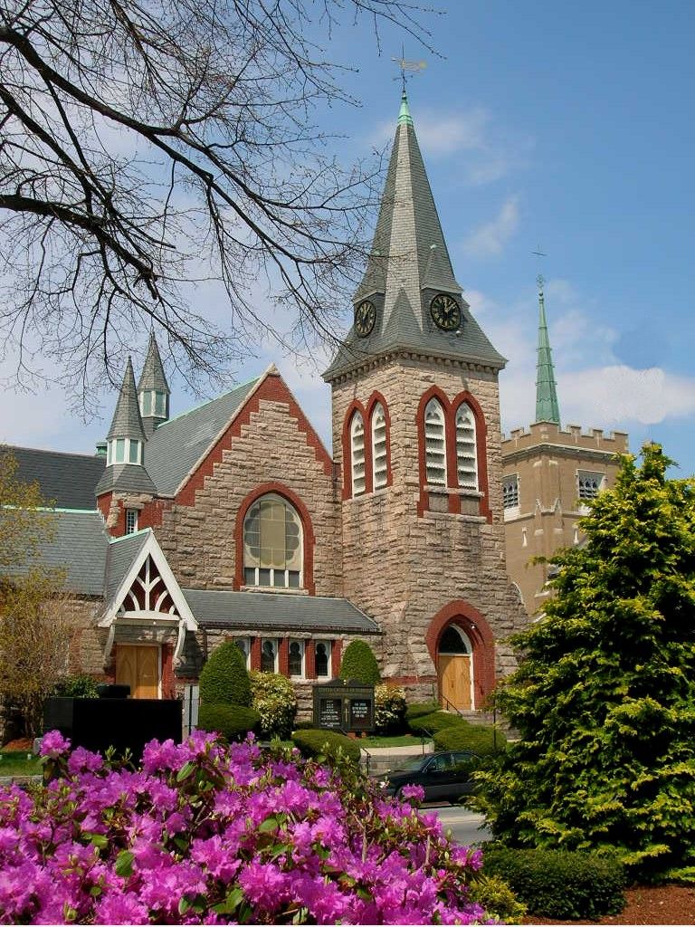Historic church in Norwood, MA featuring classic New England stone architecture with a tall clock tower, pointed steeples, and surrounding spring landscaping with blooming flowers and greenery.