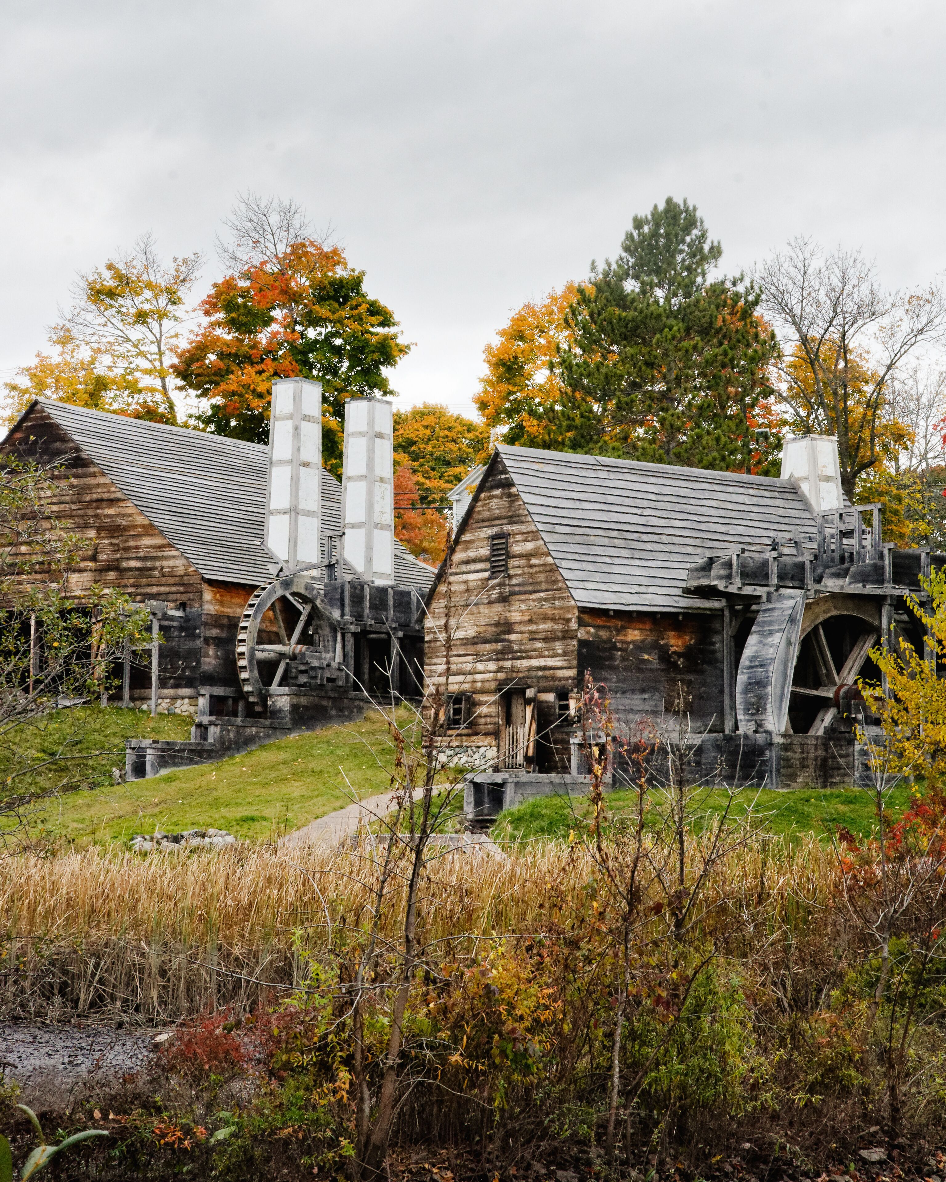 Historic water-powered iron works buildings at Saugus Iron Works National Historic Site in Saugus, MA, featuring wooden structures, large water wheels, and autumn foliage in the background