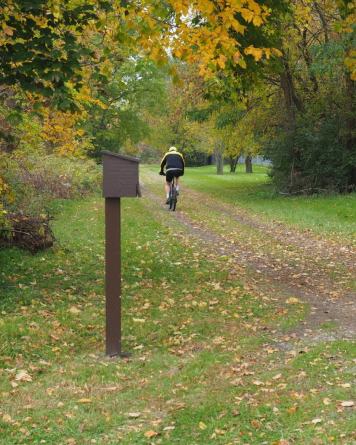 Cyclist riding along a grassy, tree-lined section of the Clinton River Trail in Pontiac, Michigan, on an overcast autumn day