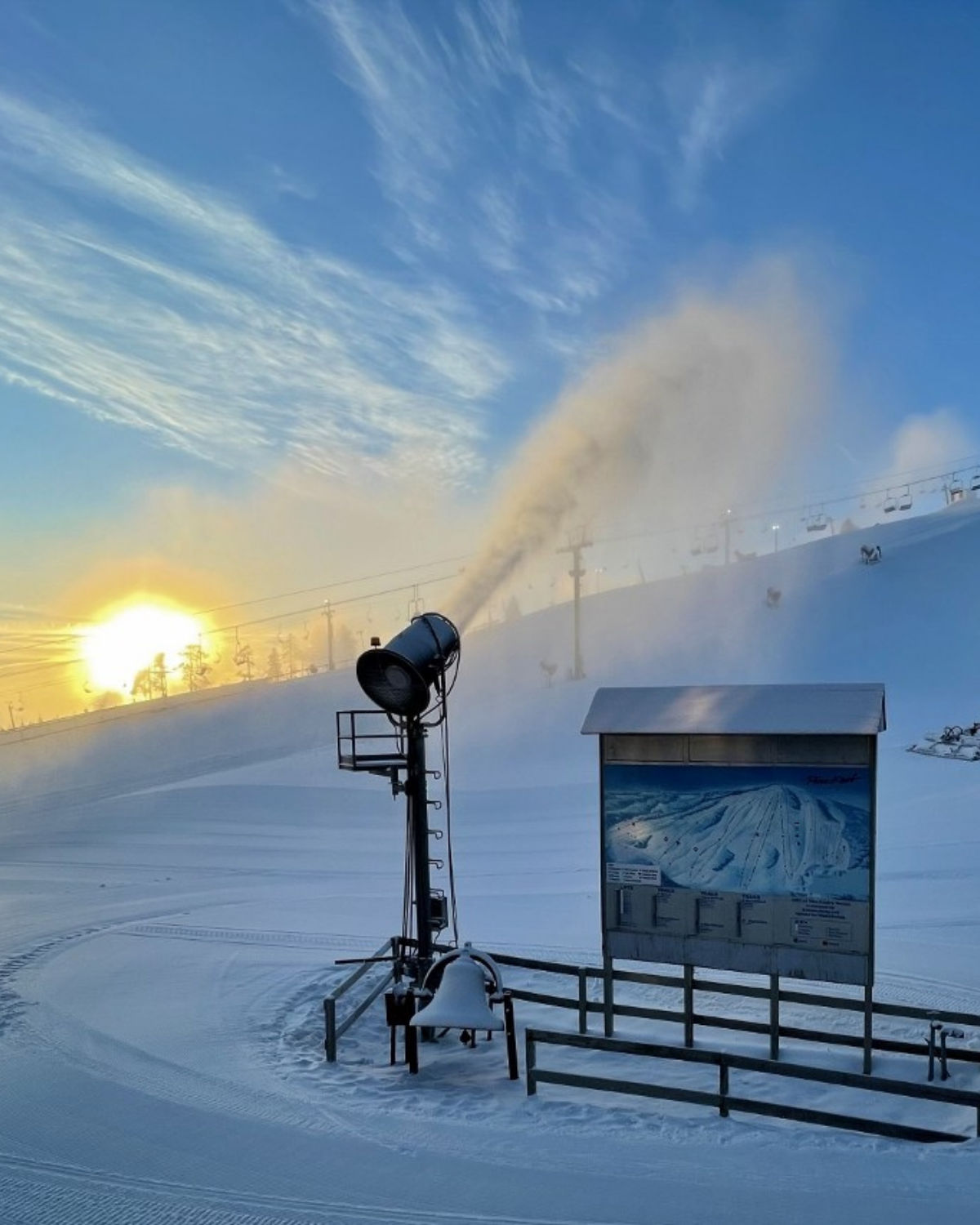 Snowy sunrise at Pine Knob Ski and Snowboard Resort in Waterford, Michigan, with a snow gun blowing fresh powder across a groomed slope beside a large trail map sign