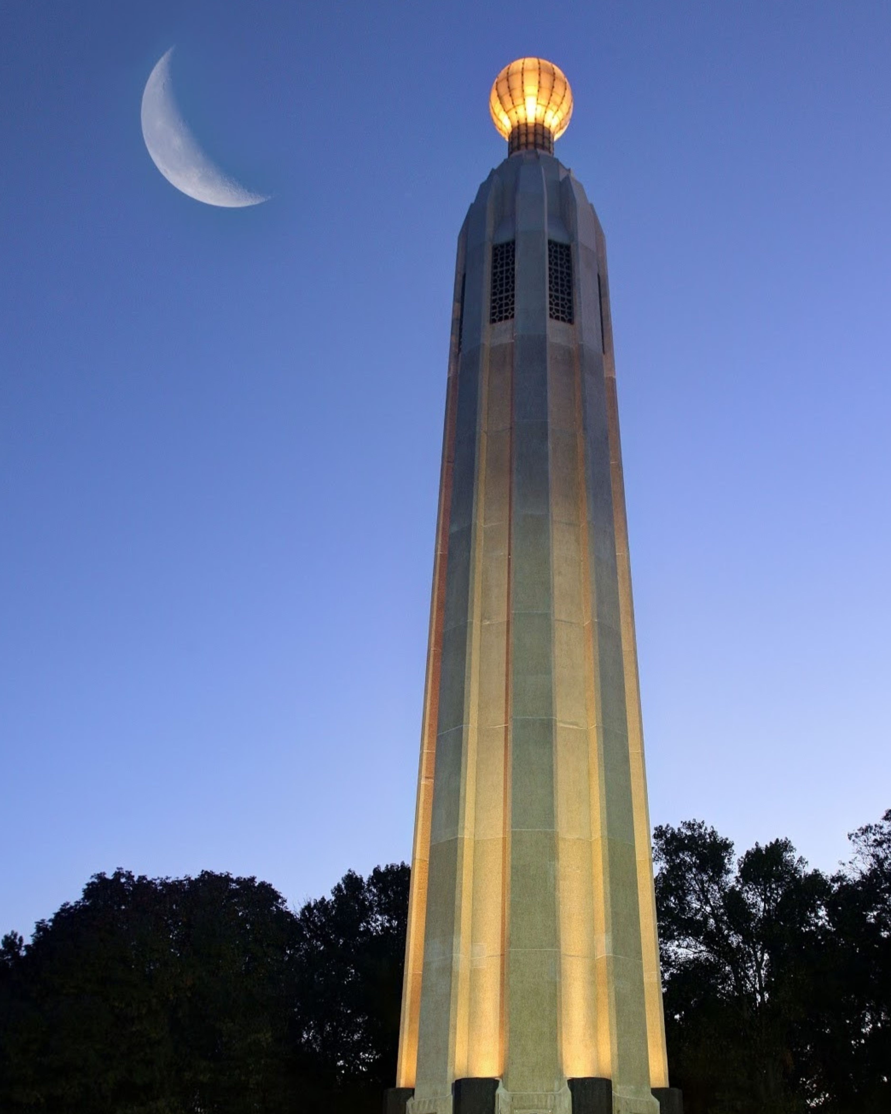 The Thomas Alva Edison Memorial Tower in Edison, New Jersey, illuminated at dusk beneath a crescent moon. The iconic monument rises above surrounding trees, honoring inventor Thomas Edison and marking the site of his historic Menlo Park laboratory.