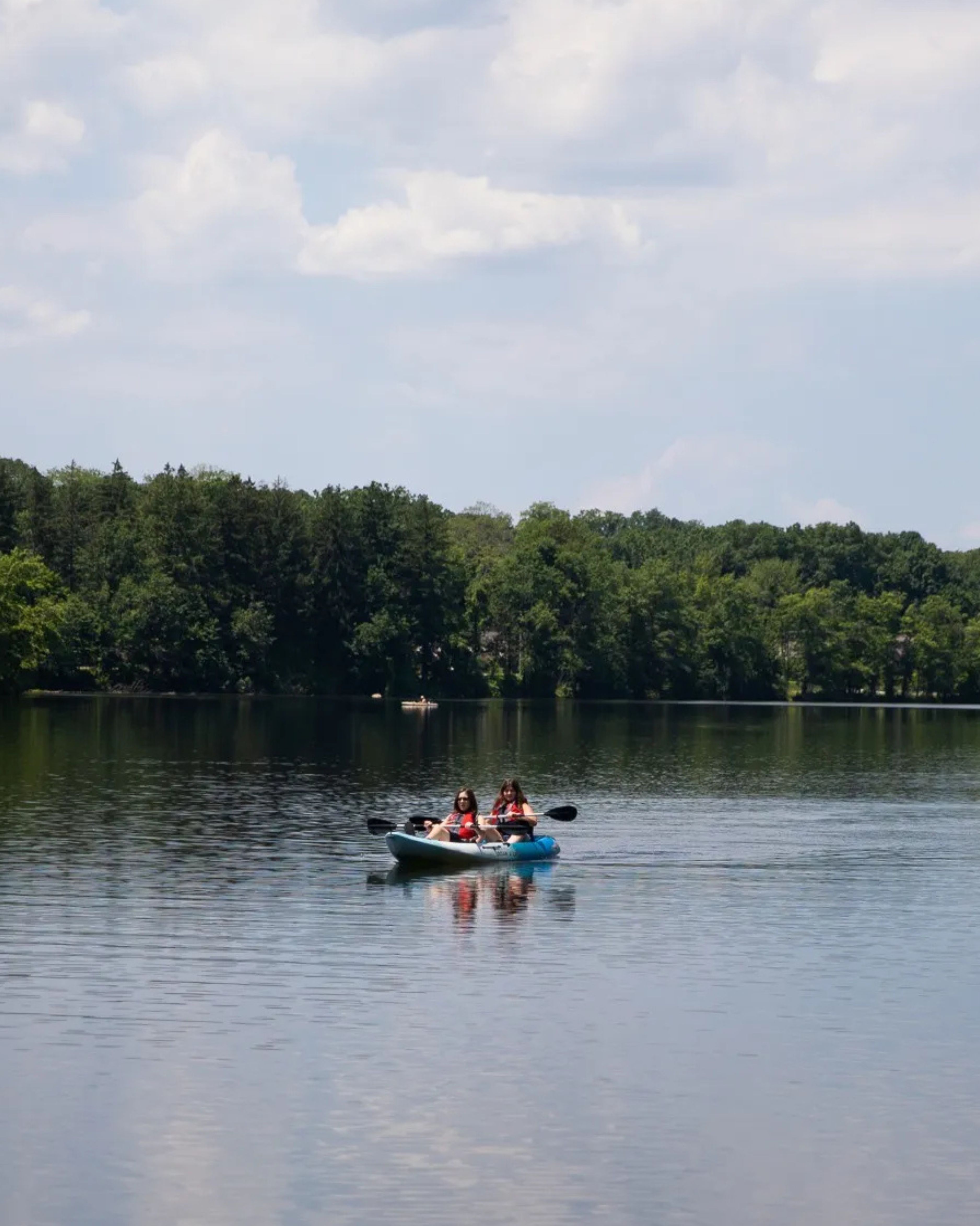 Scenic view of a lake in Franklin Lakes, New Jersey, with two people kayaking on calm water surrounded by wooded shoreline under a partly cloudy sky.