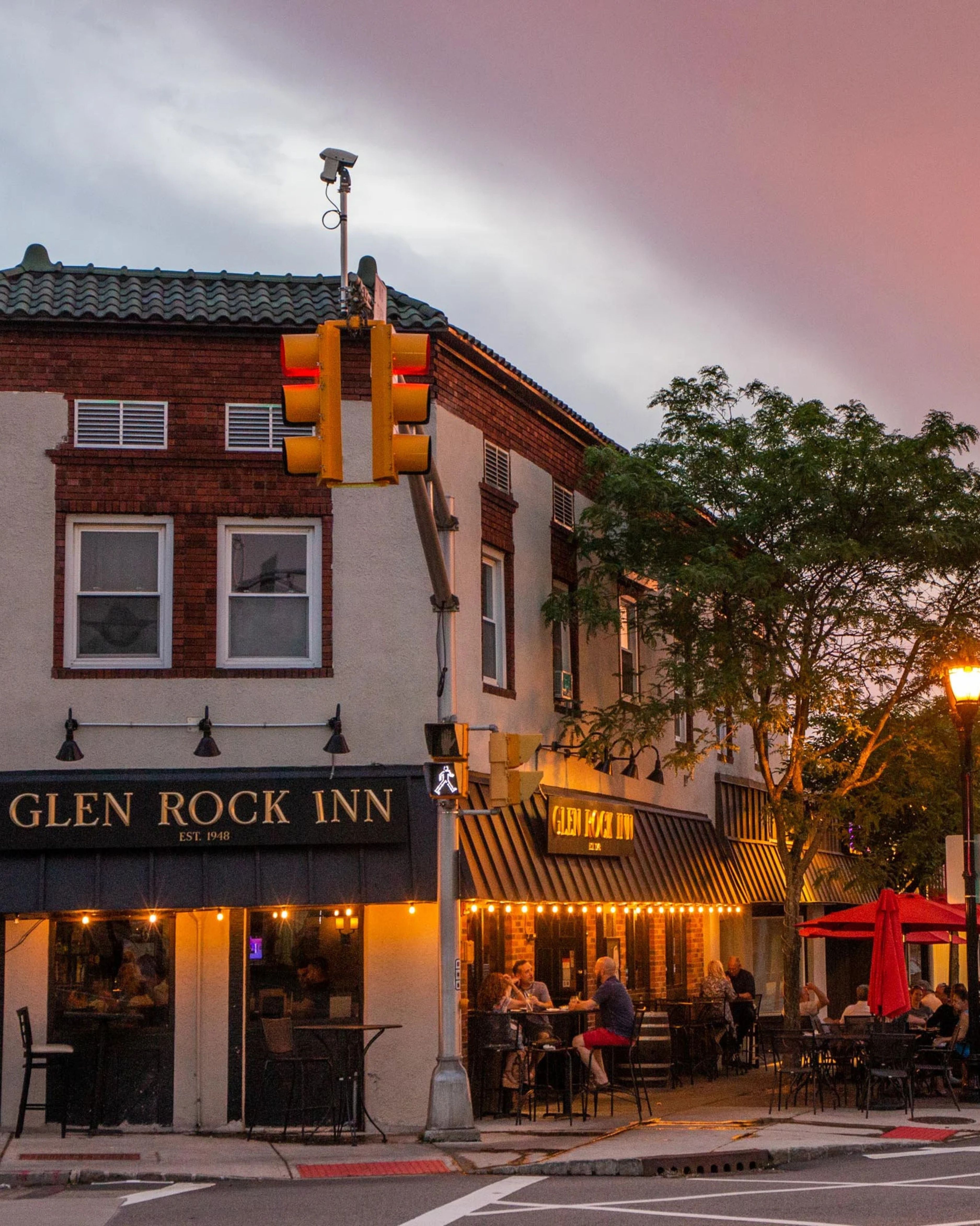 Evening street view of downtown Glen Rock, New Jersey, featuring the Glen Rock Inn restaurant with outdoor dining, glowing storefront lights, pedestrians gathered outside, and a neighborhood intersection beneath a colorful sunset sky.