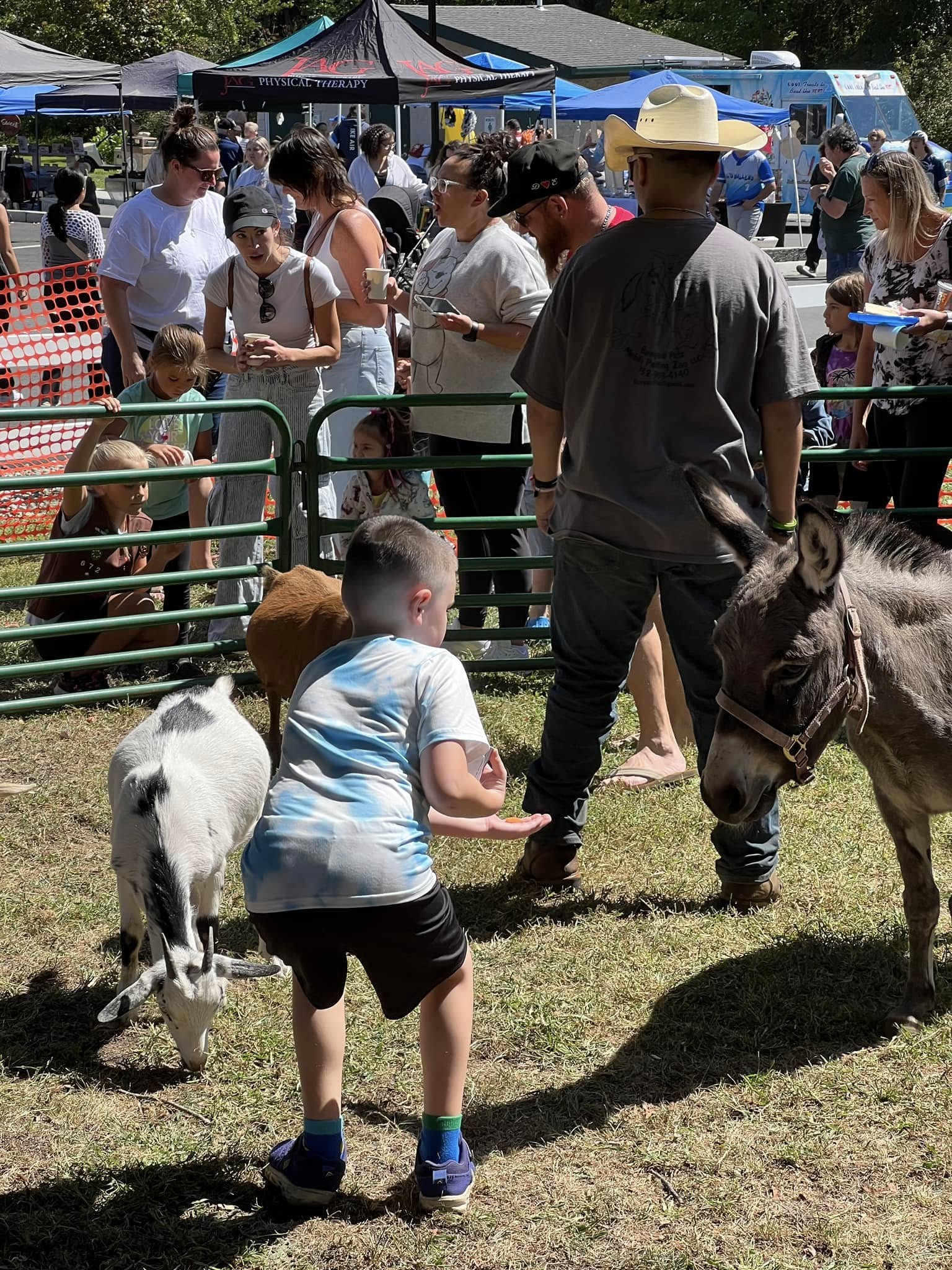 Families visiting goats and a donkey at an outdoor petting zoo and community event in Hazlet, New Jersey, with local vendors and activities in the background.