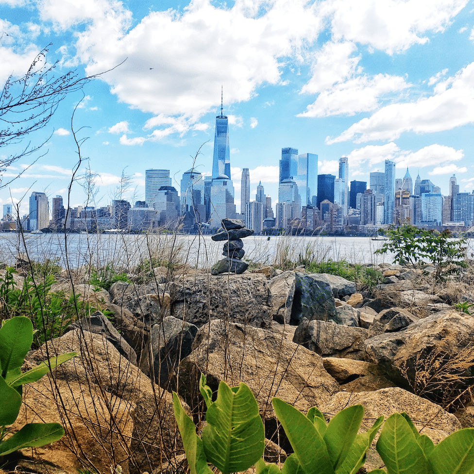 A scenic view of the Manhattan skyline from Jersey City’s waterfront, featuring One World Trade Center in the distance, rocky shoreline in the foreground, and a small stacked stone cairn overlooking the Hudson River under a bright, partly cloudy sky.