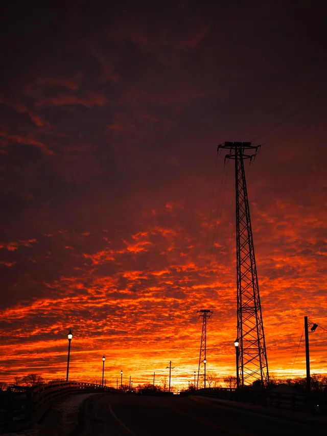 Dramatic sunset over Keyport, New Jersey, featuring silhouetted utility towers, streetlights, and a pedestrian bridge beneath a vibrant orange and red evening sky.
