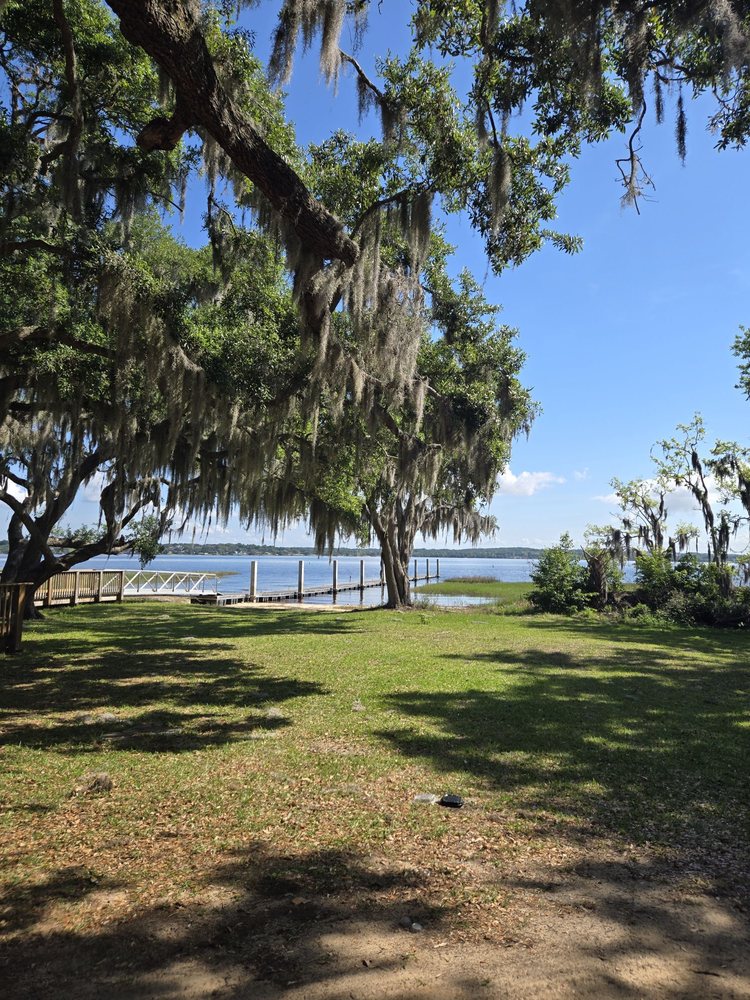 Waterfront park along the shores of Lake Hiawatha, New Jersey, featuring mature oak trees draped with Spanish moss, a grassy shoreline, and a wooden dock extending into the calm water. The peaceful setting offers scenic views of the lake beneath a clear blue sky.