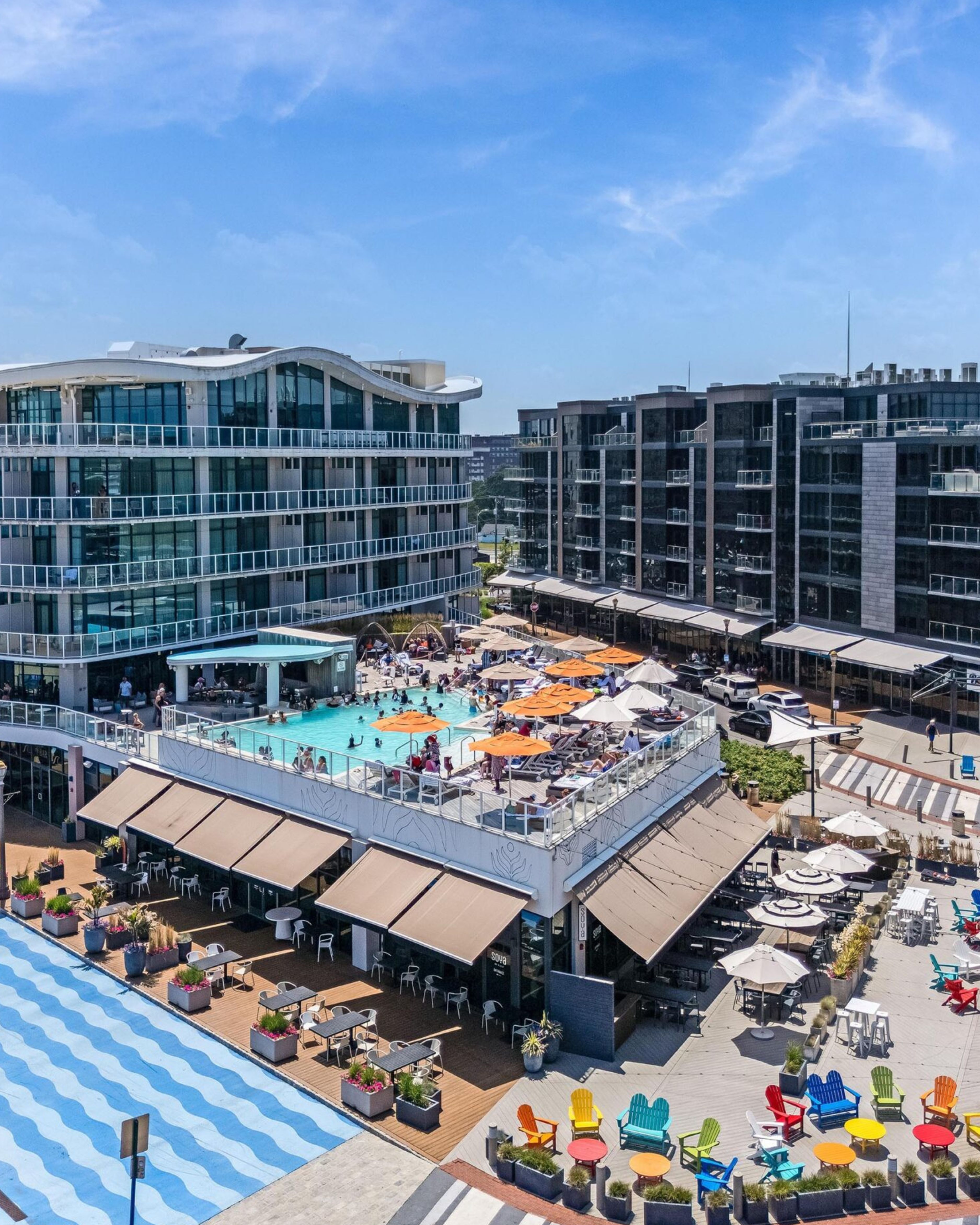 Aerial view of Pier Village in Long Branch, New Jersey, featuring modern waterfront residences, a rooftop swimming pool, restaurants, outdoor dining areas, colorful Adirondack chairs, and pedestrian gathering spaces.
