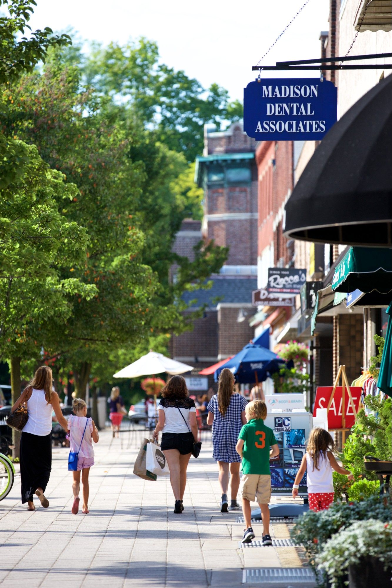 Tree-lined downtown street in Madison, NJ with families walking along a busy sidewalk, local storefronts and signage visible, including a “Madison Dental Associates” sign, capturing a lively and walkable small-town atmosphere.